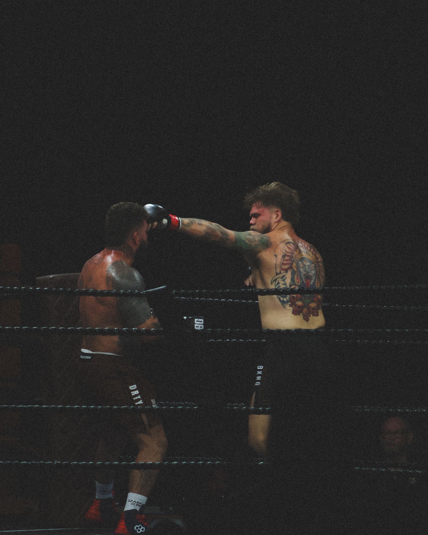 Two boxers exchange punches in a dimly lit boxing ring during an intense match.
