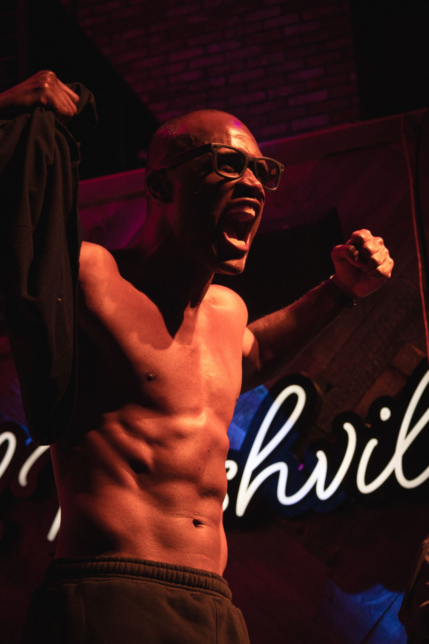 Shirtless performer passionately singing on stage with neon signage in background.