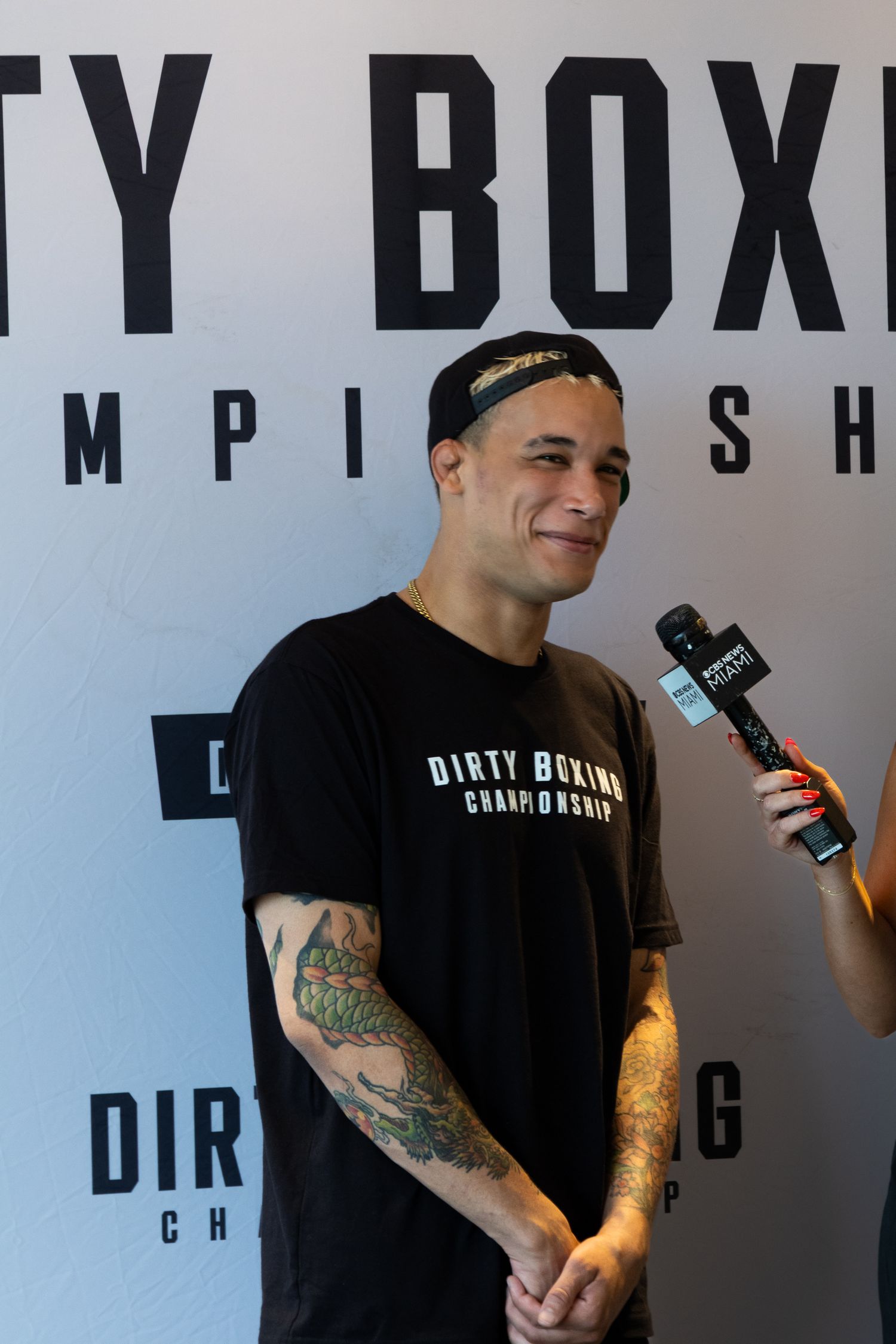 Person in black shirt with tattoos smiles during interview at boxing event.