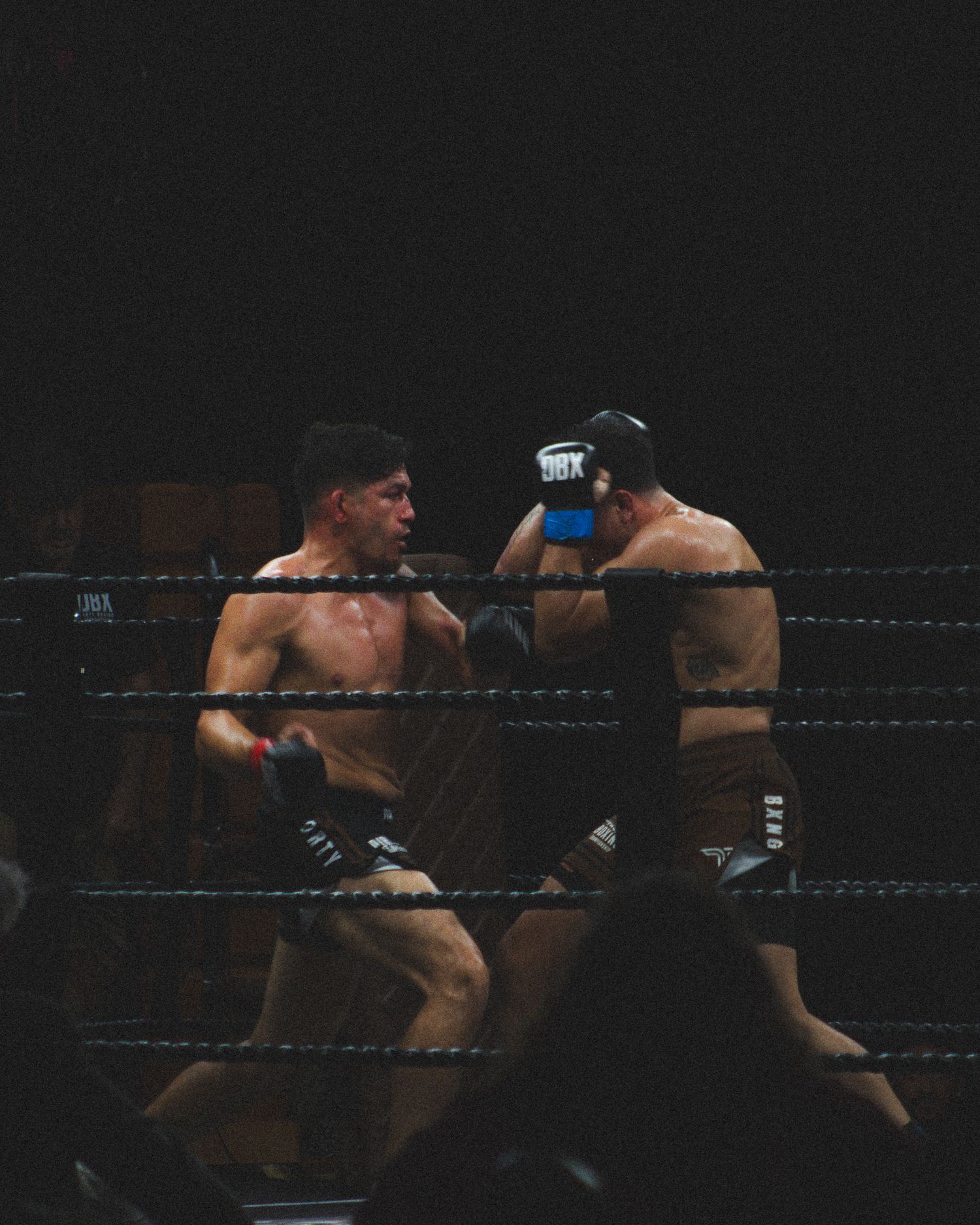 Fighters trade punches in a dimly lit boxing ring during an intense match.