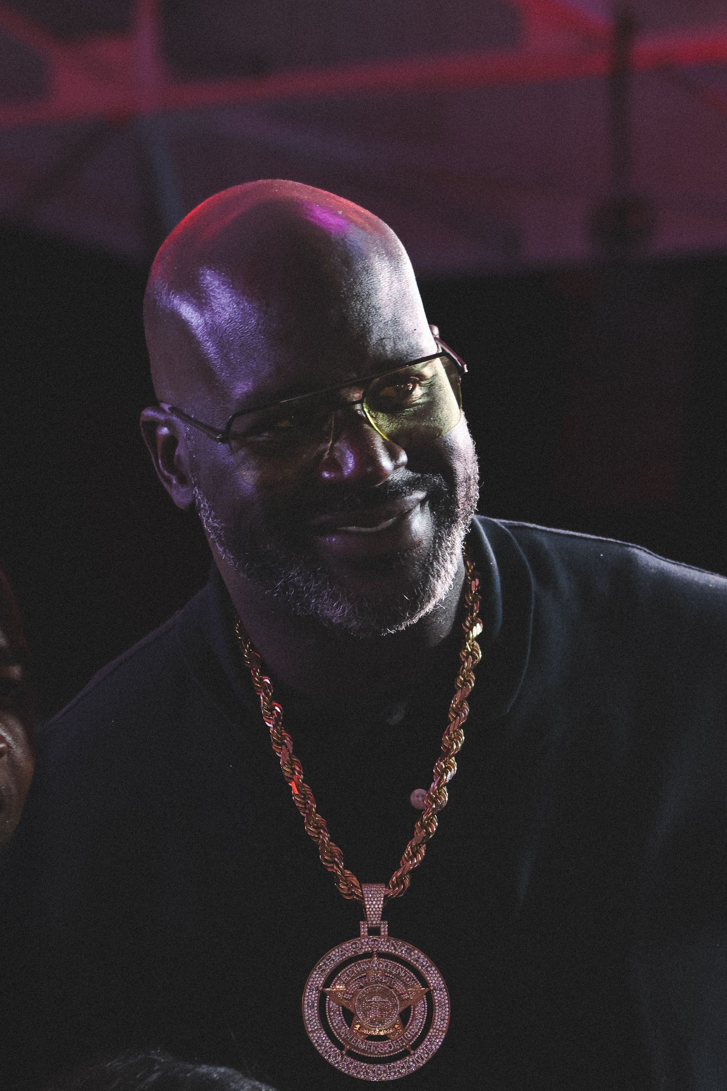 A man wearing glasses and a gold medallion necklace smiles under purple lighting.
Shaq