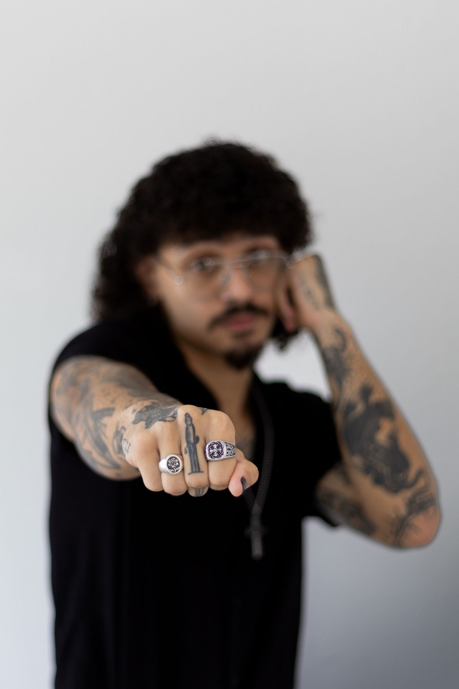 A tattooed person with long dark hair extends their hand showing rings toward the camera.