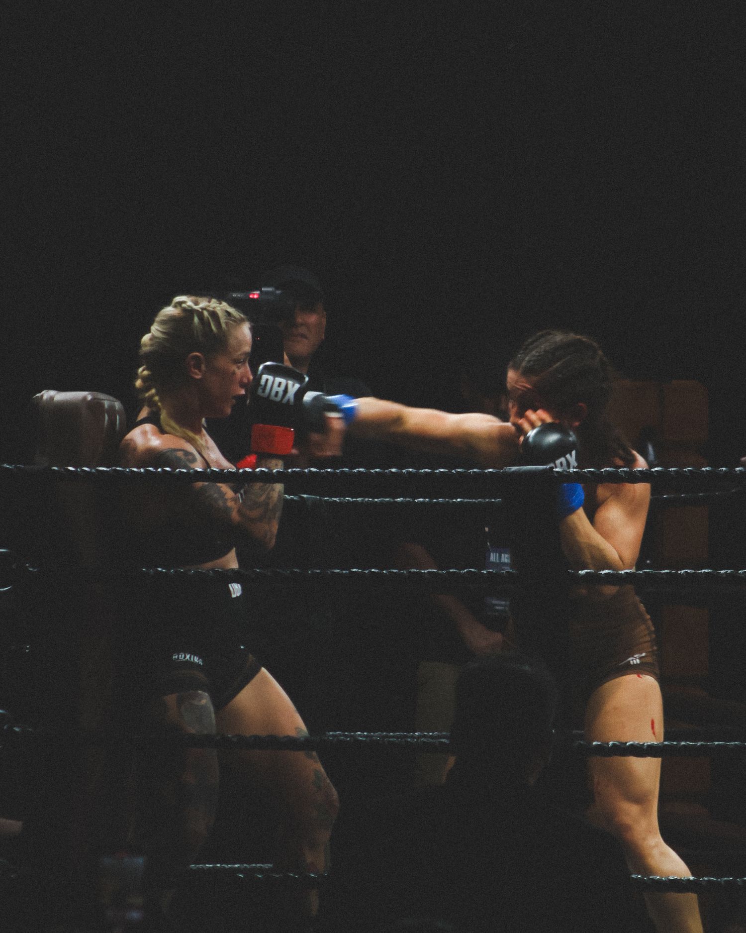 Two boxers exchange punches in a darkly lit boxing ring during an intense match.