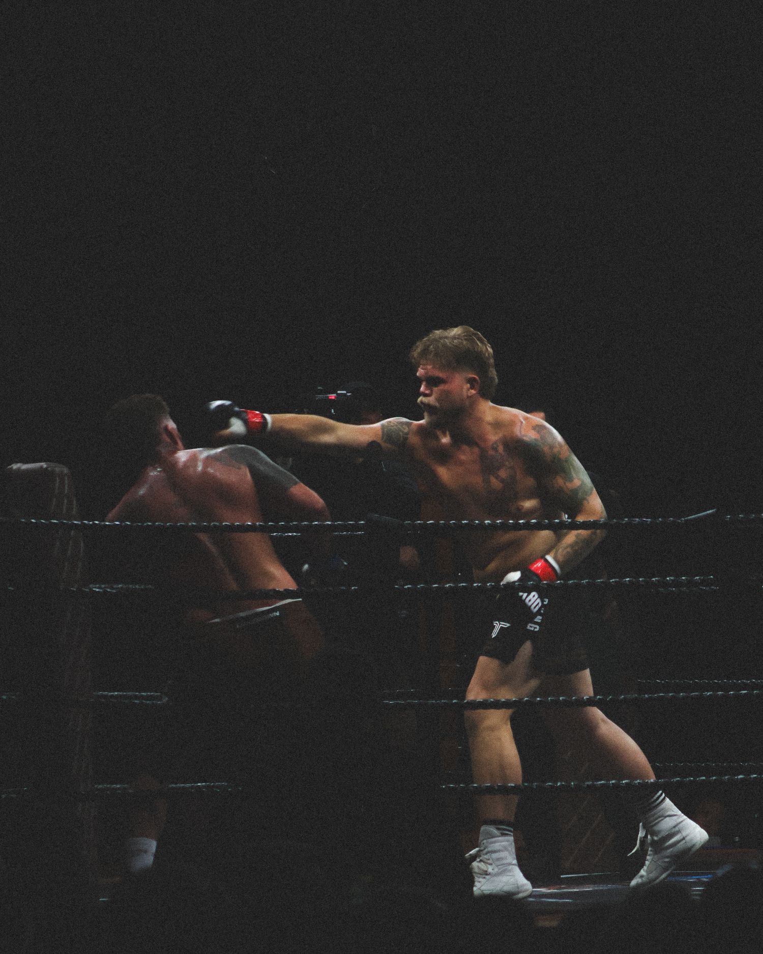 Boxers engage in a fierce exchange near the ropes of a darkened boxing ring.