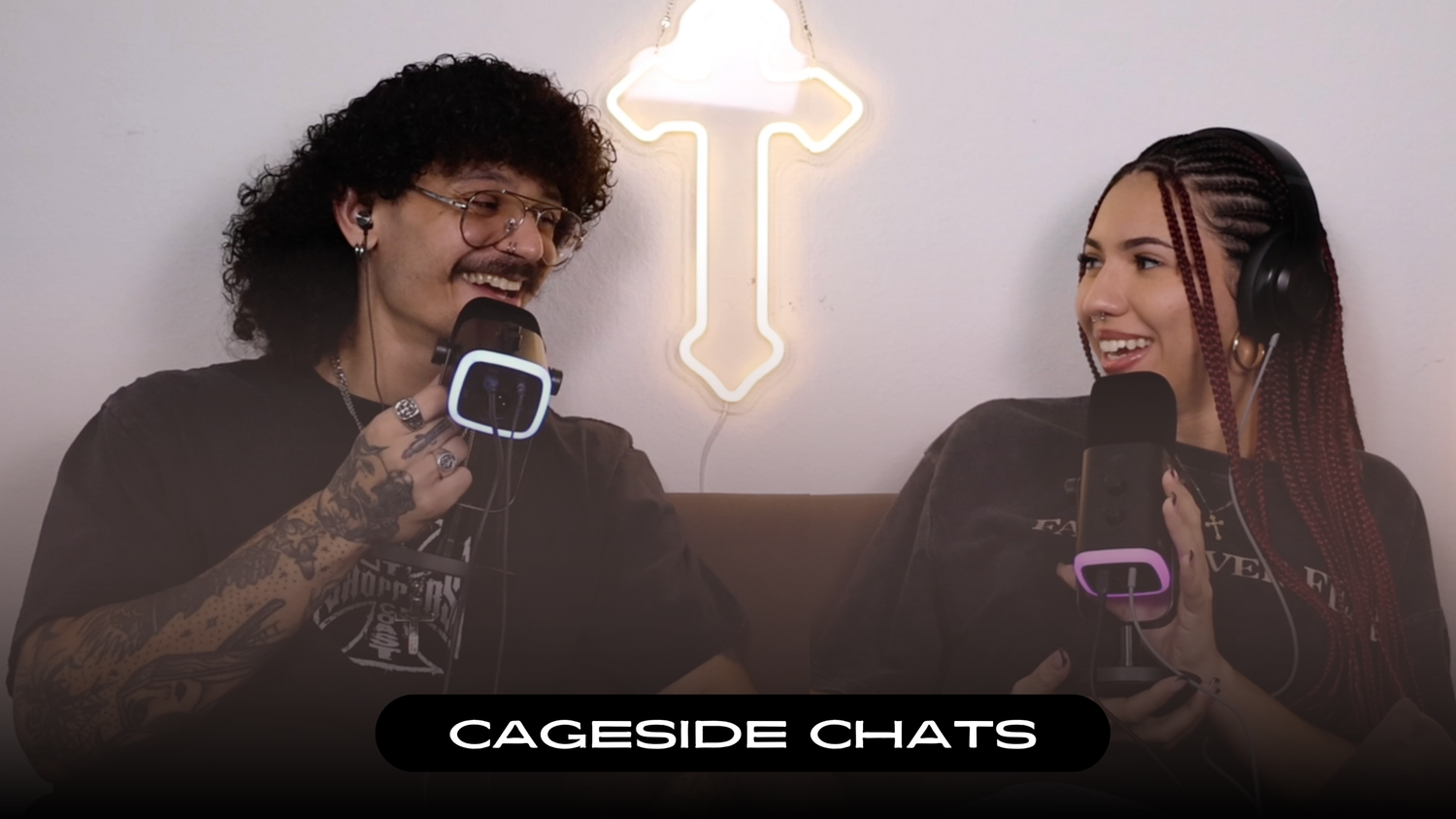 Two people having a conversation in front of a glowing neon sword sign during a Cageside Chats interview.