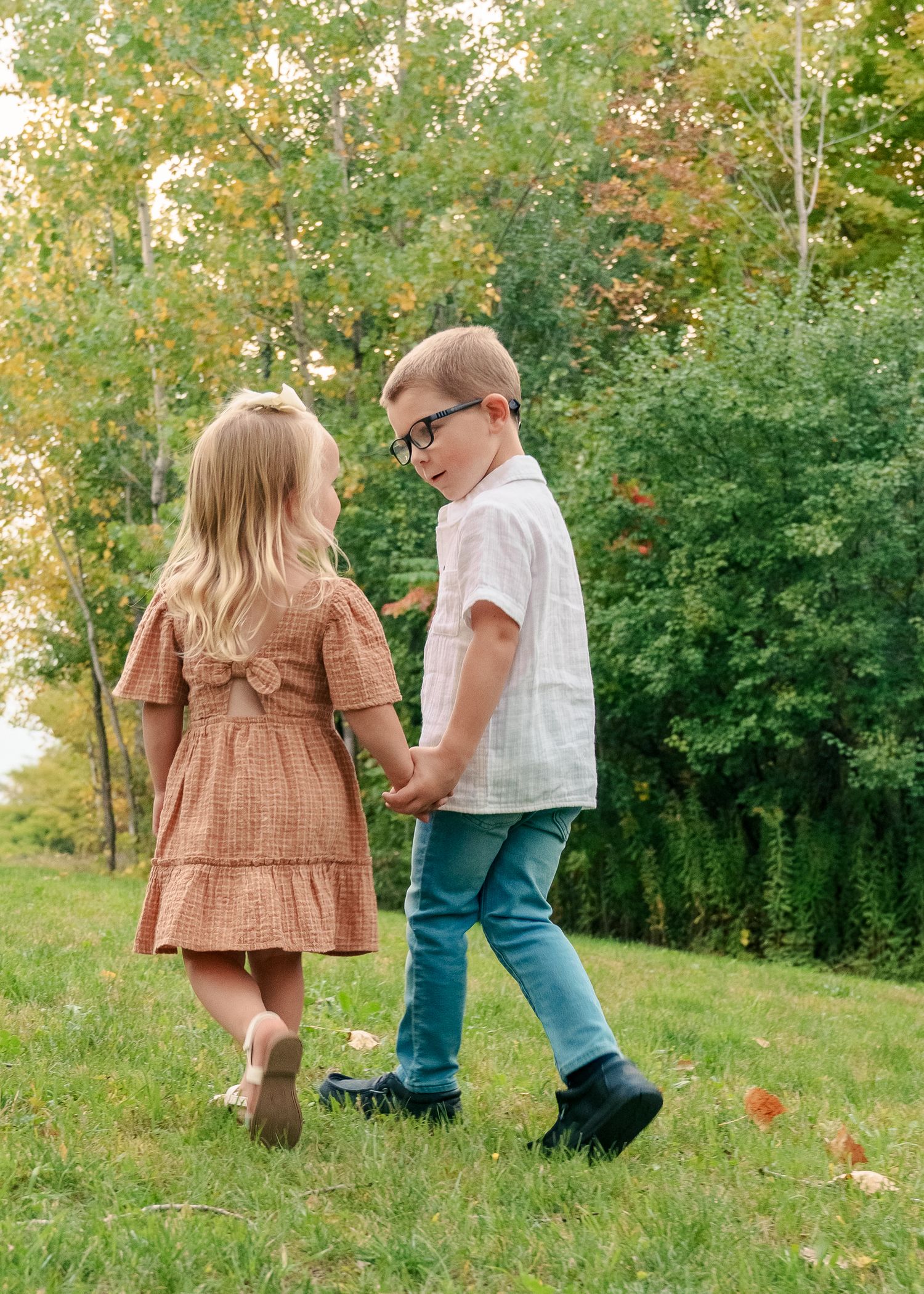 Two young children walking together on a grassy hill with autumn trees in the background.