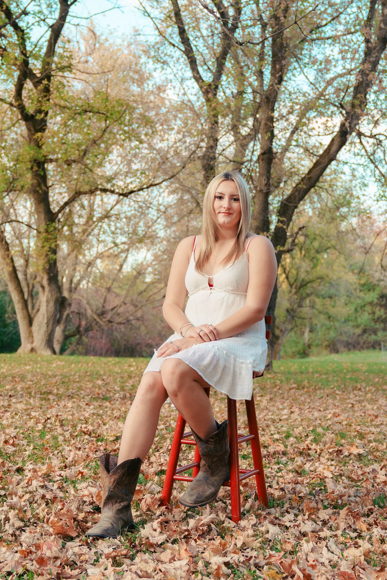 A person in a white dress sits on a red stool surrounded by autumn leaves in a wooded outdoor setting.