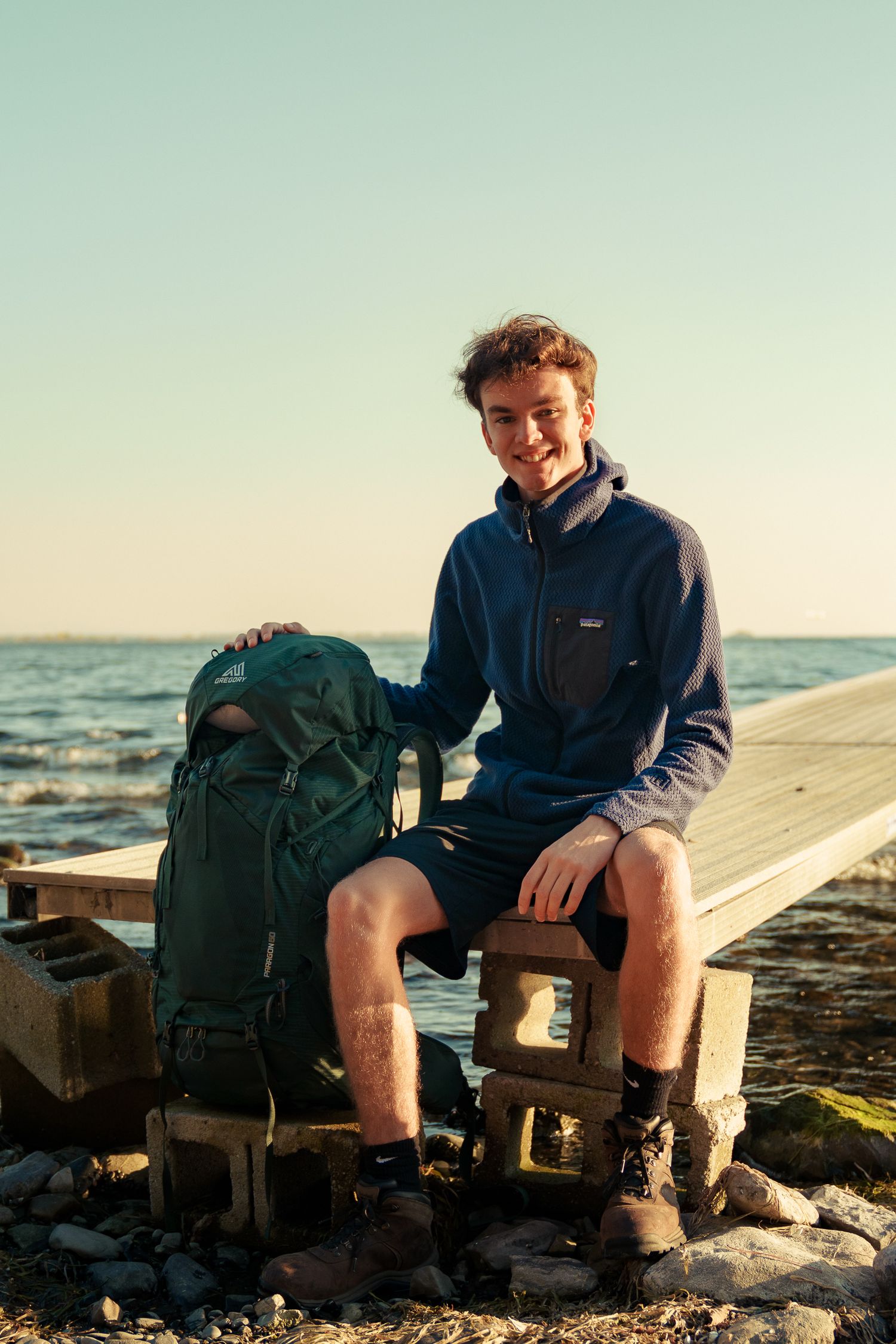 Person wearing blue jacket sits with backpack on rocky beach at sunset with ocean waves in background.