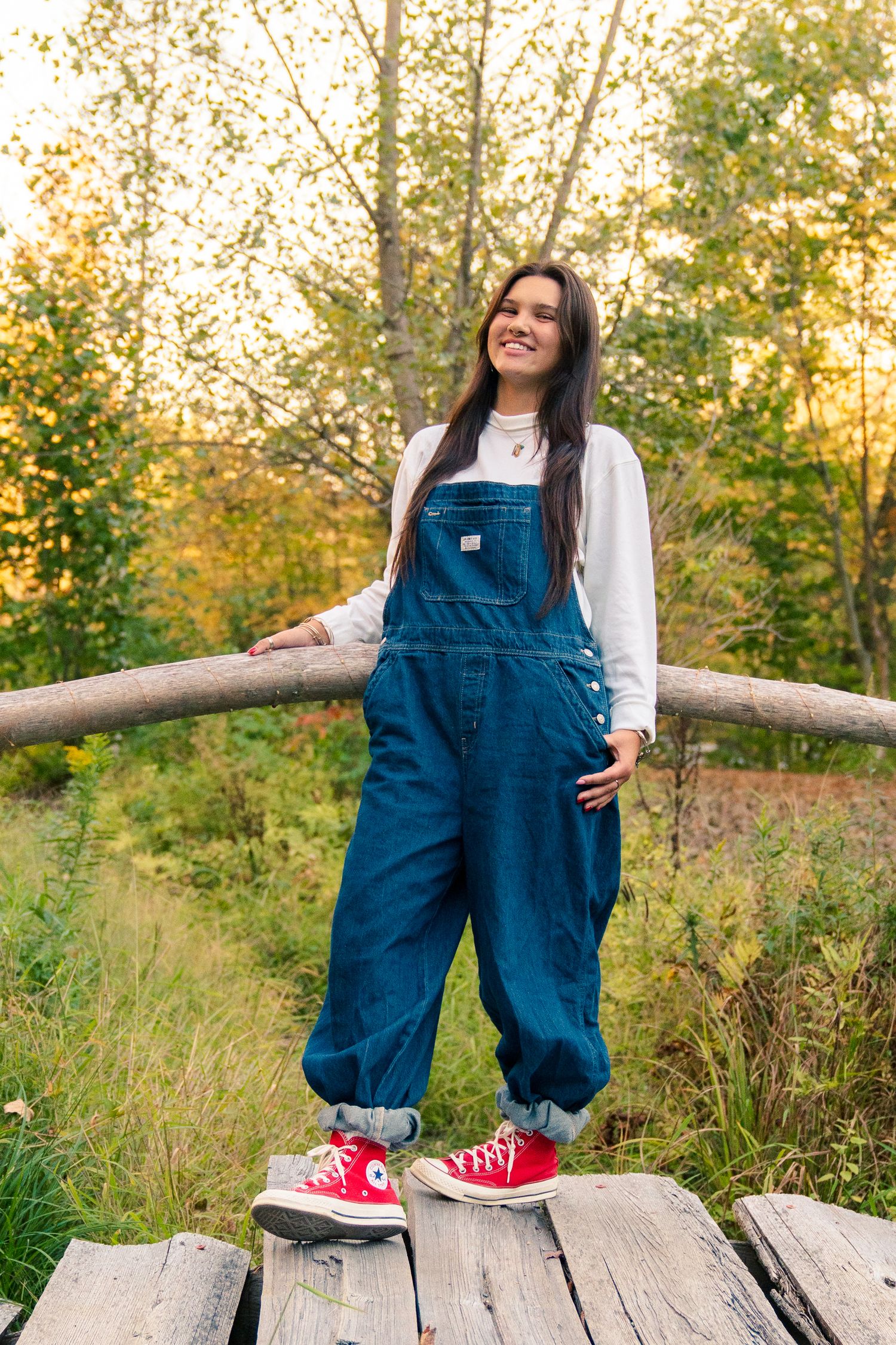 Maternity photo series showing denim overalls and red sneakers against autumn foliage at sunset.