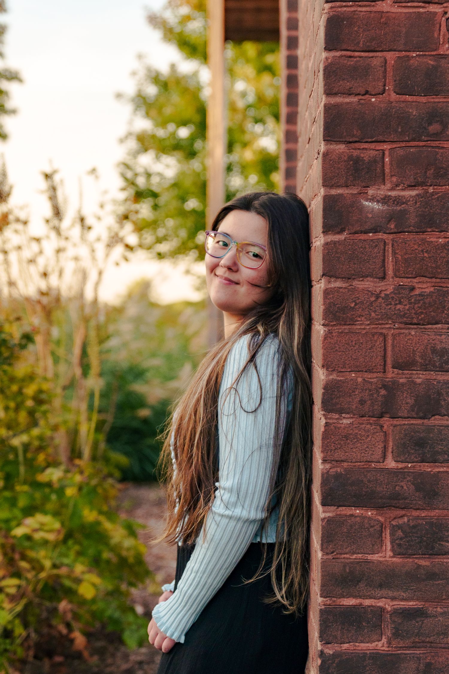 Portrait of someone in a white knit sweater and glasses leaning against a brick wall at sunset.