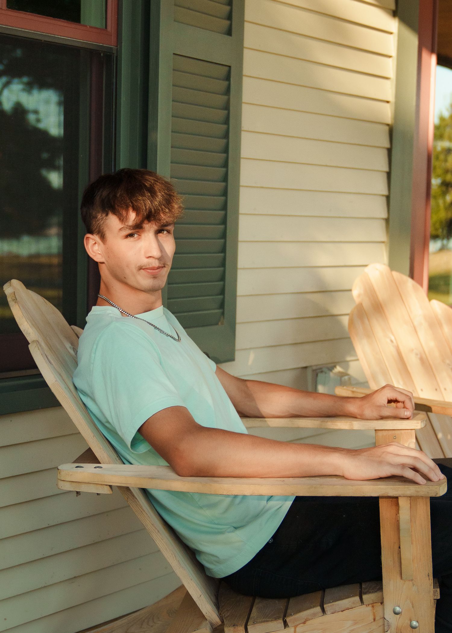 Relaxed poses in a mint t-shirt while sitting in an Adirondack chair on a porch.