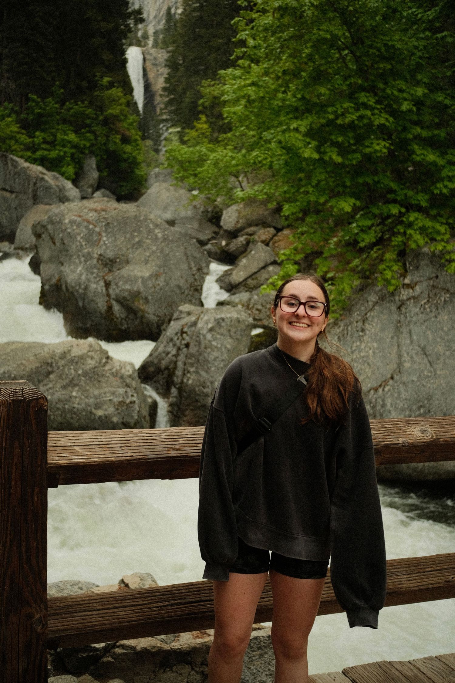 Person in black sits on wooden railing near rocky waterfall in lush forest setting.