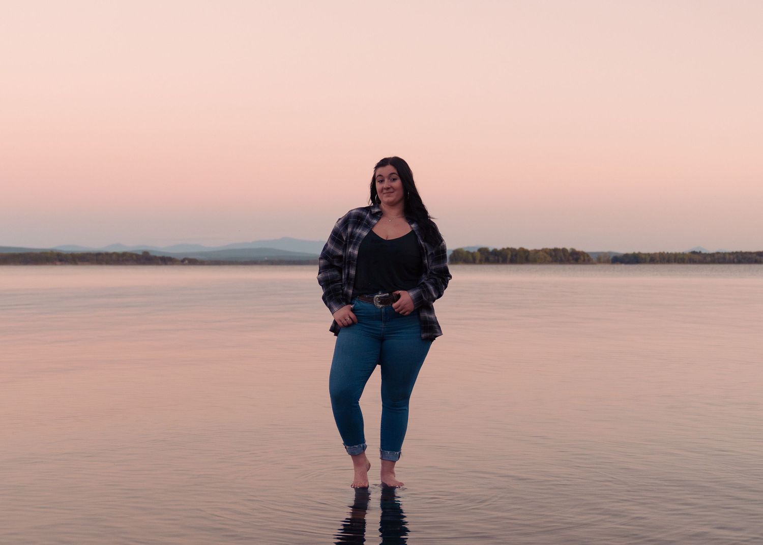 A figure stands on a beach at sunset wearing a black jacket and blue jeans against a pink-hued sky and calm water.