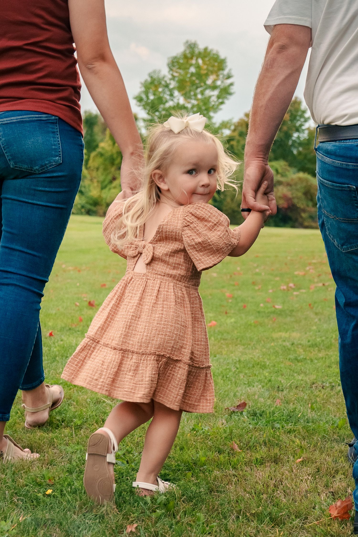 A series of photos showing a toddler in a peach-colored dress walking on grass while holding hands with adults.