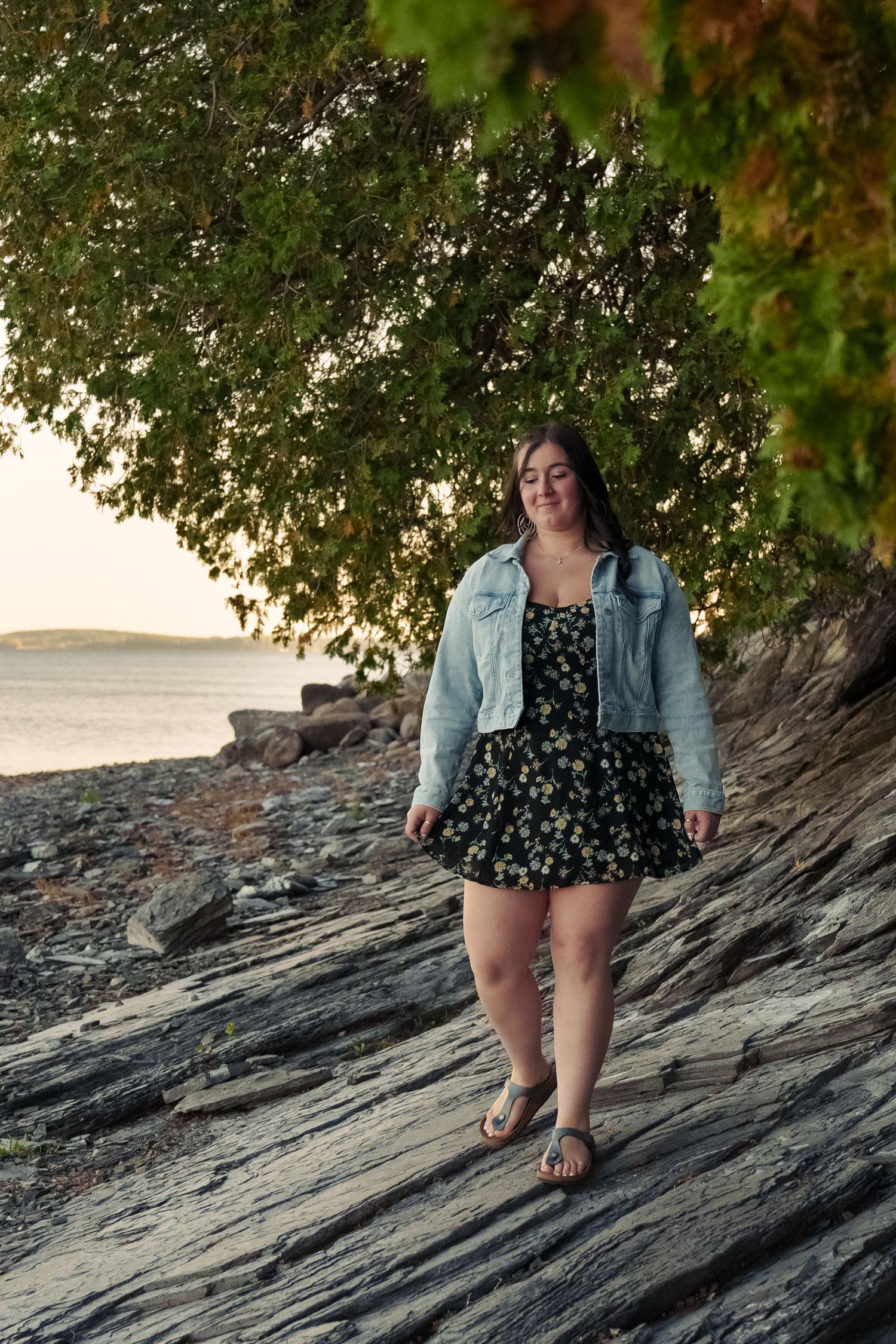 Person in denim jacket and floral dress sits on rocky coastal outcrop at sunset with trees overhead.