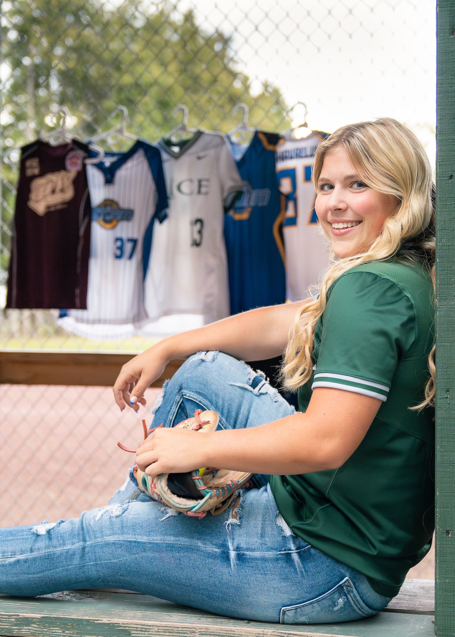 Person in green shirt and ripped jeans sits casually on ground with sports jerseys displayed on wall behind them.