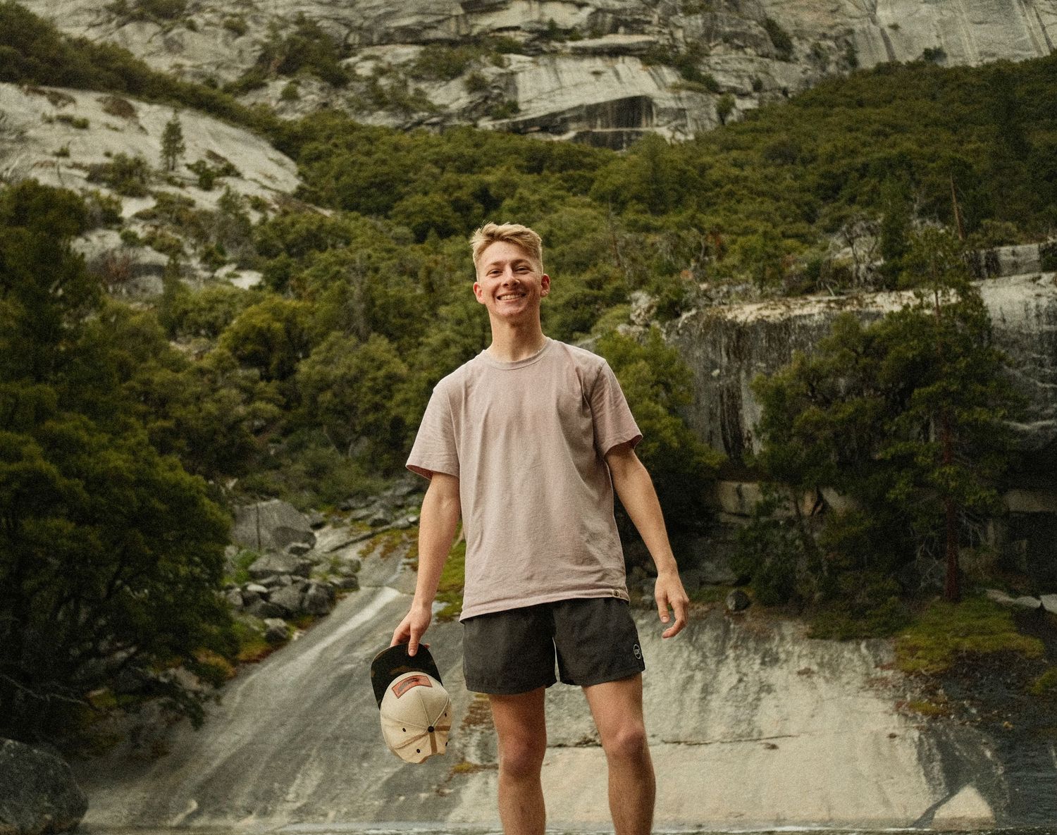 A person stands on rocky terrain against dramatic mountain cliffs wearing casual hiking attire.