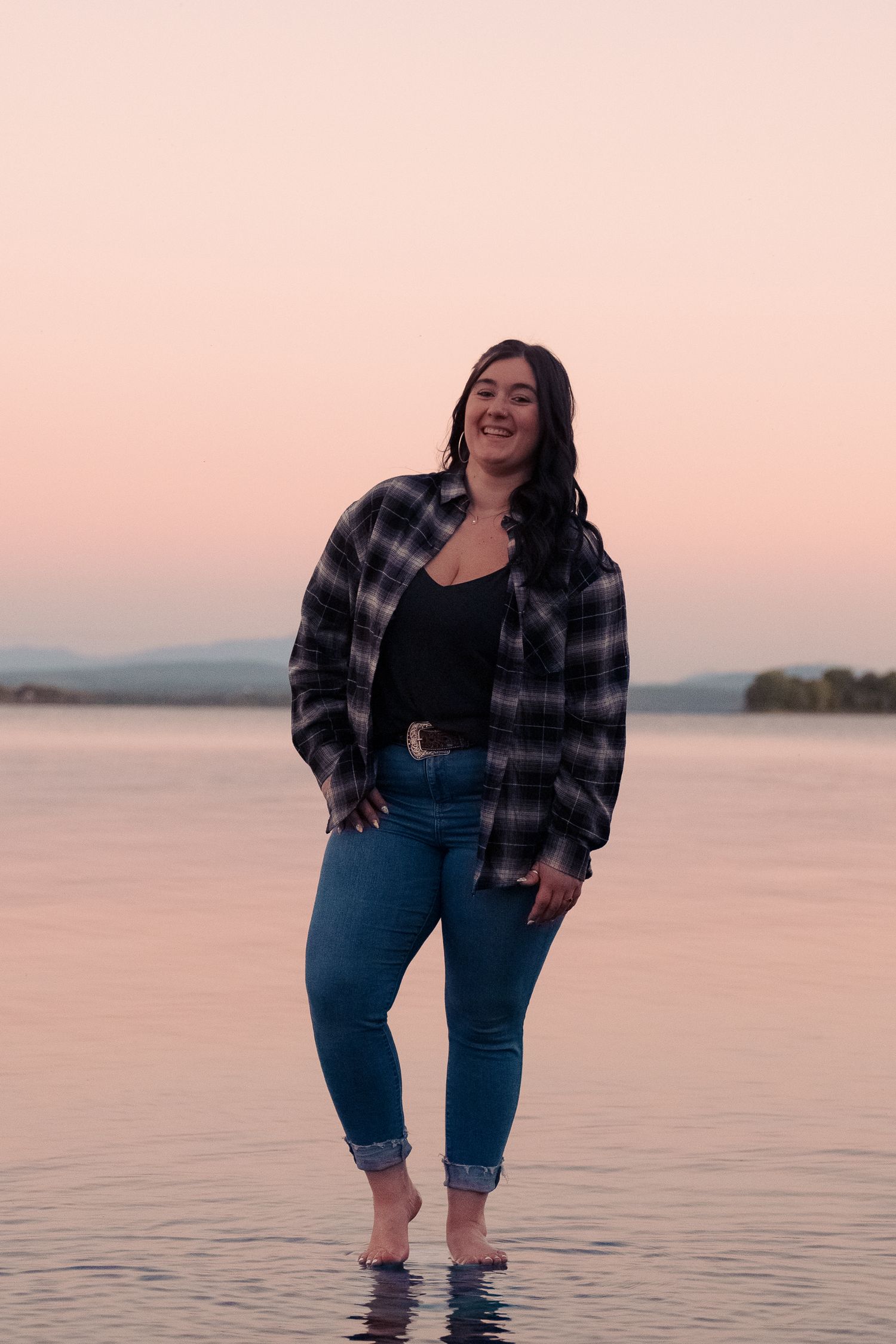 A woman in plaid flannel and jeans poses playfully in shallow lake water during sunset.