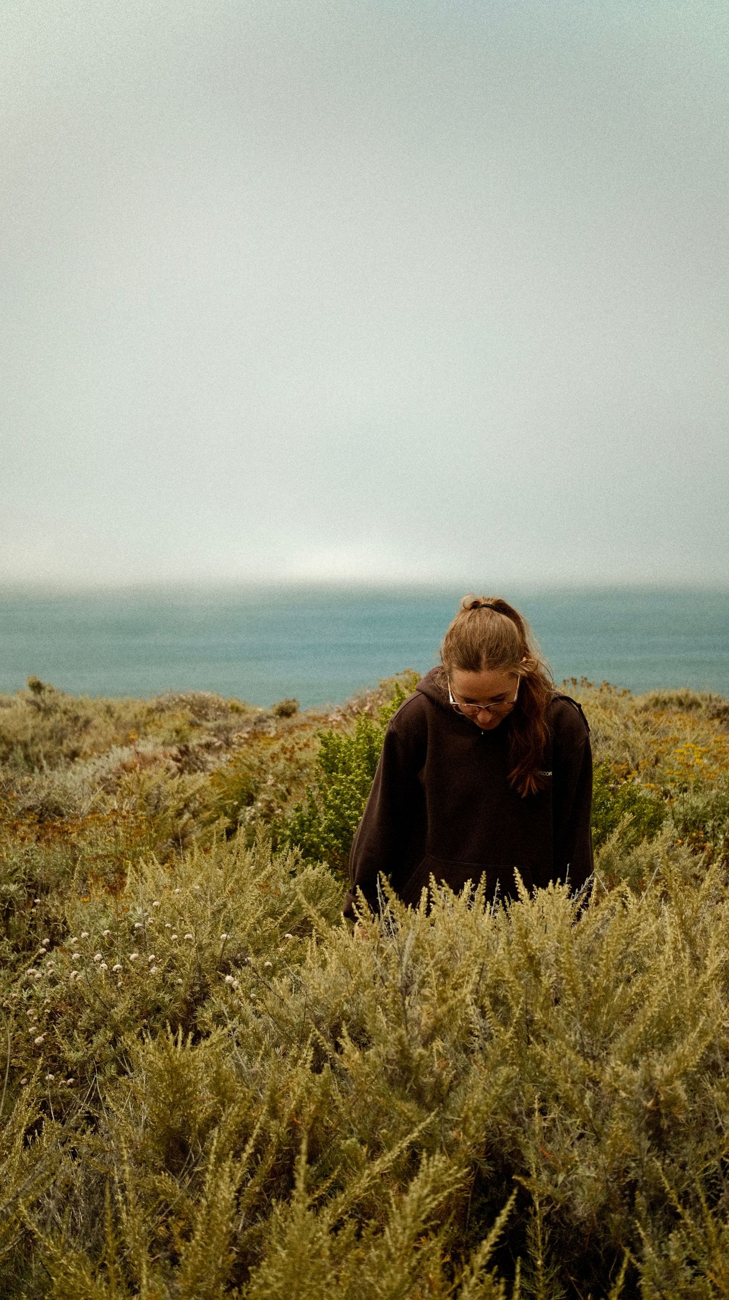 Figure in dark clothing crouches among coastal shrubs with ocean vista in background.