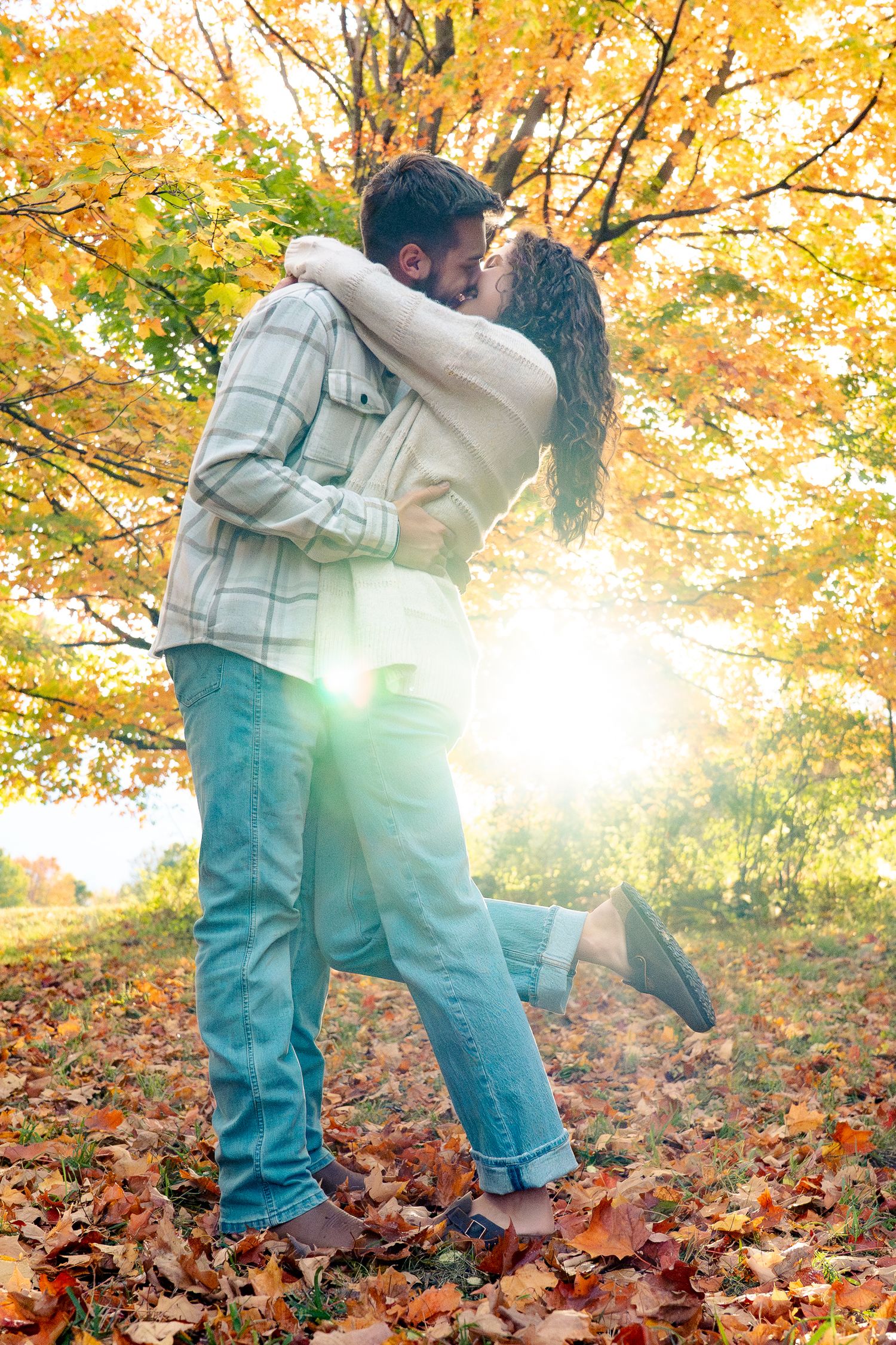 A couple embraces romantically in a park surrounded by golden autumn foliage and fallen leaves on the ground.