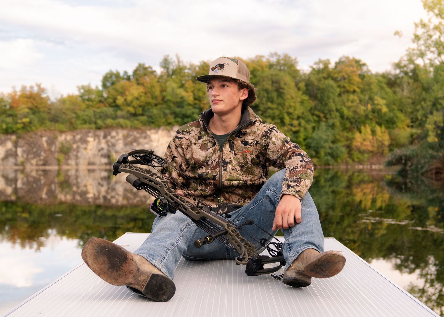 A hunter in camouflage gear sits on a dock by a serene lake during autumn.