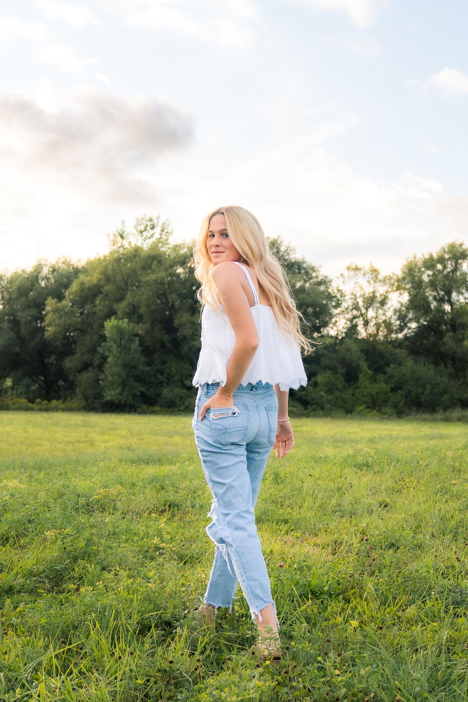 A person in a white off-shoulder top and light blue jeans poses in a grassy field at sunset with trees in the background.