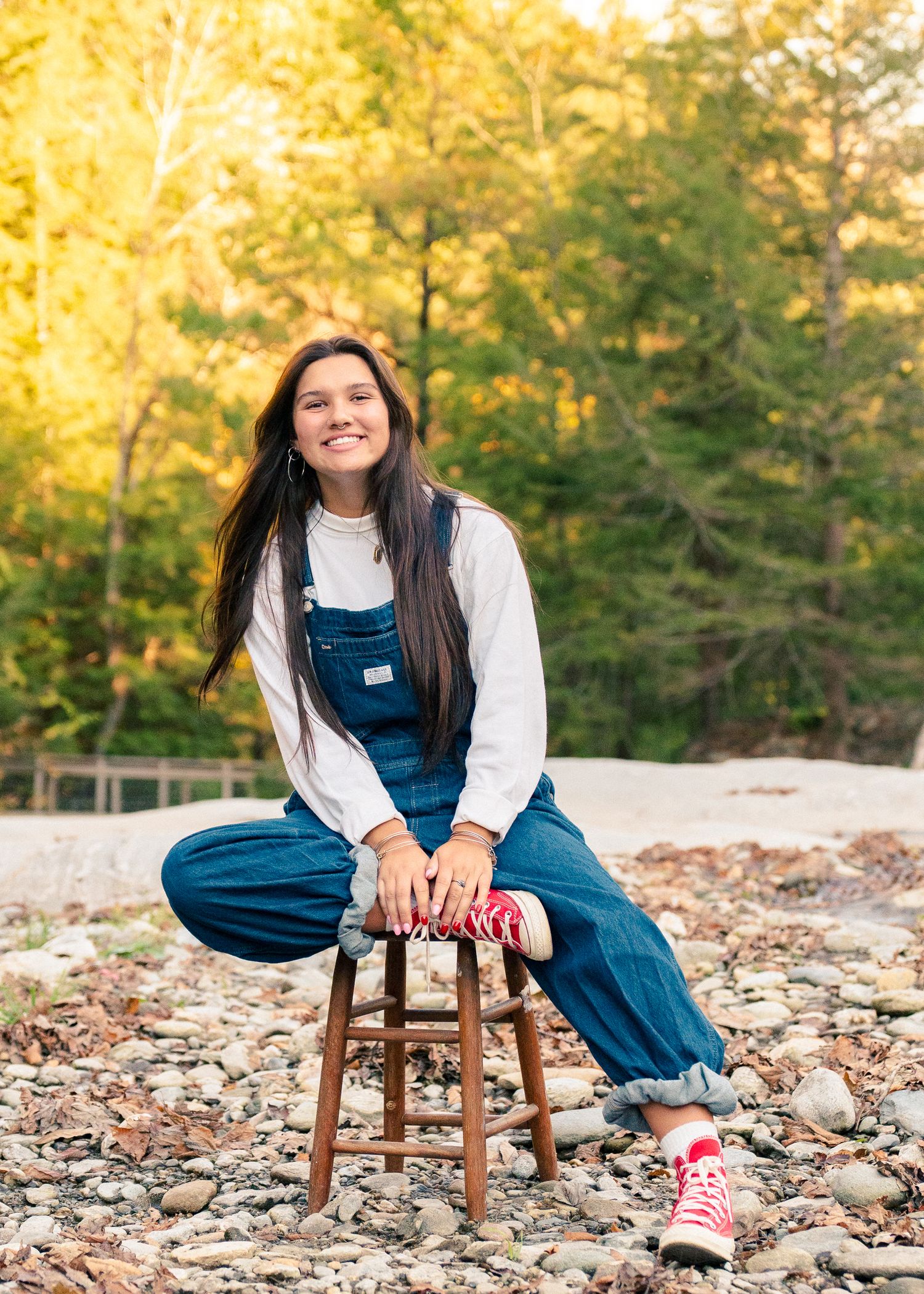 Person in blue denim overalls and white top sits on wooden stool against autumn forest backdrop.