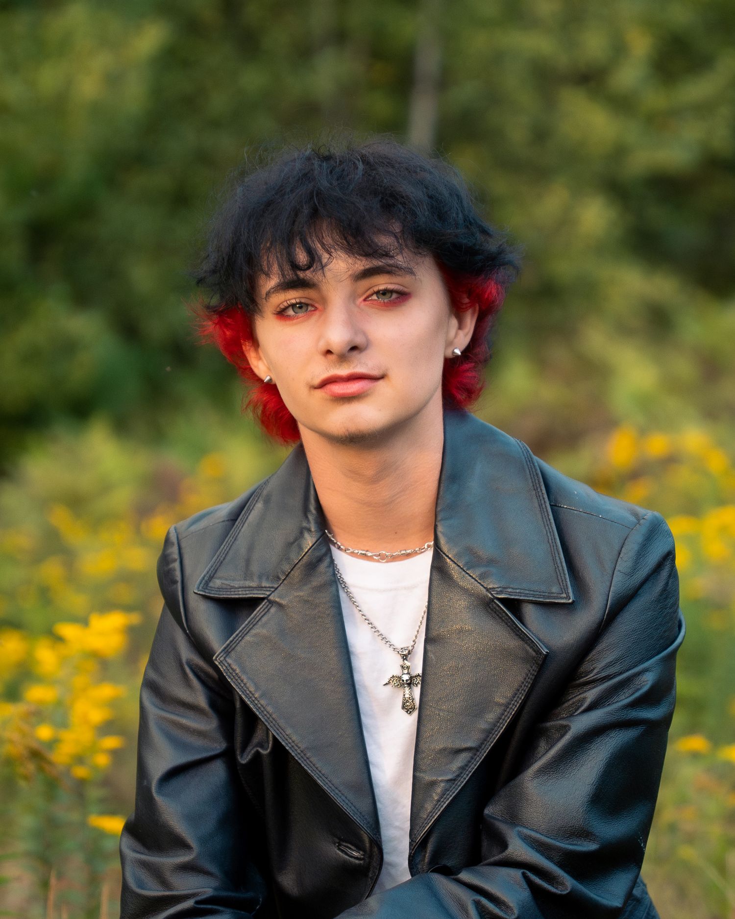 A figure in a black leather jacket and white shirt poses outdoors against a natural background with yellow flowers.