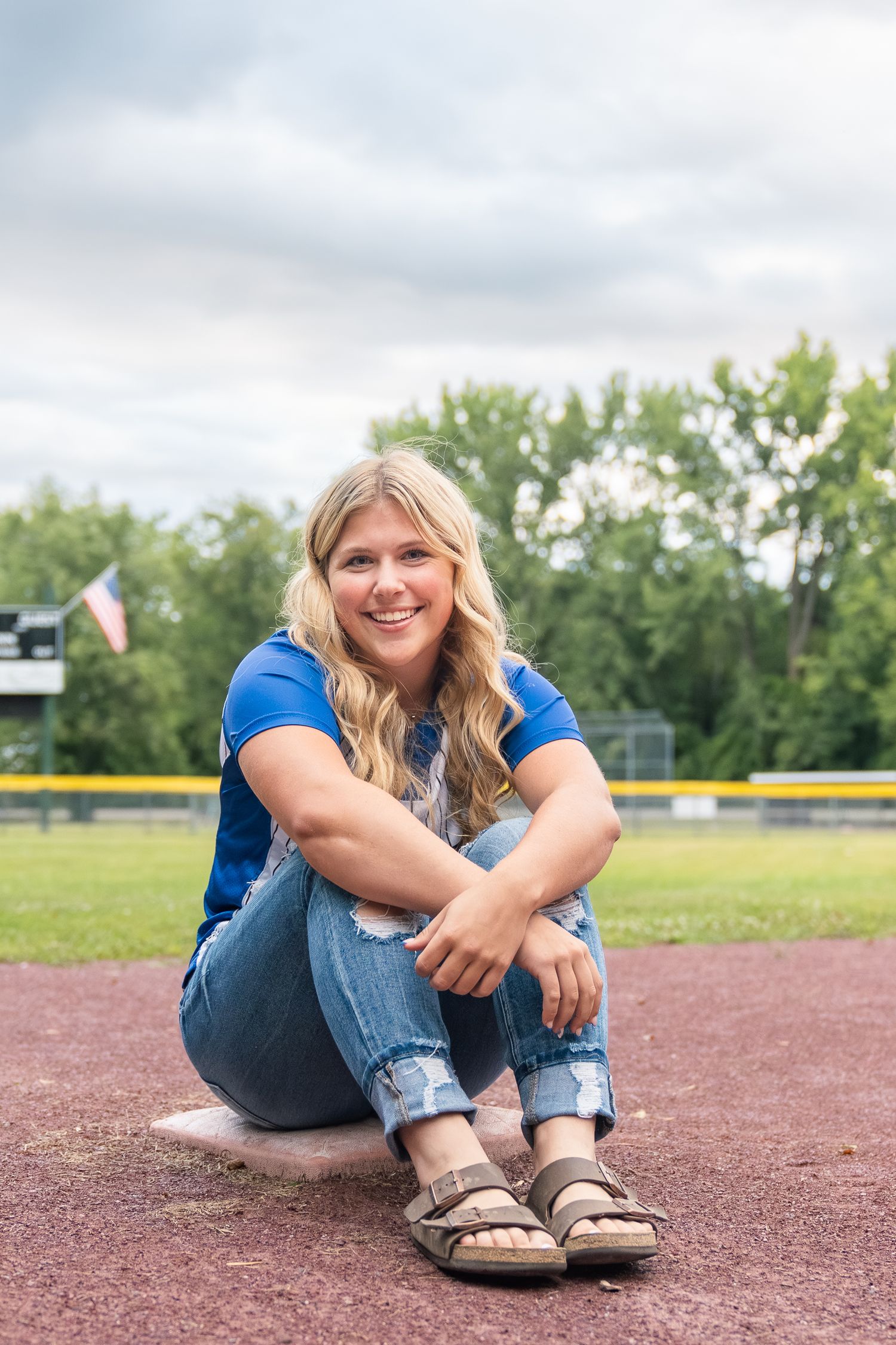 A casual summer portrait at a baseball field showing someone seated on the ground wearing a blue shirt and jeans.