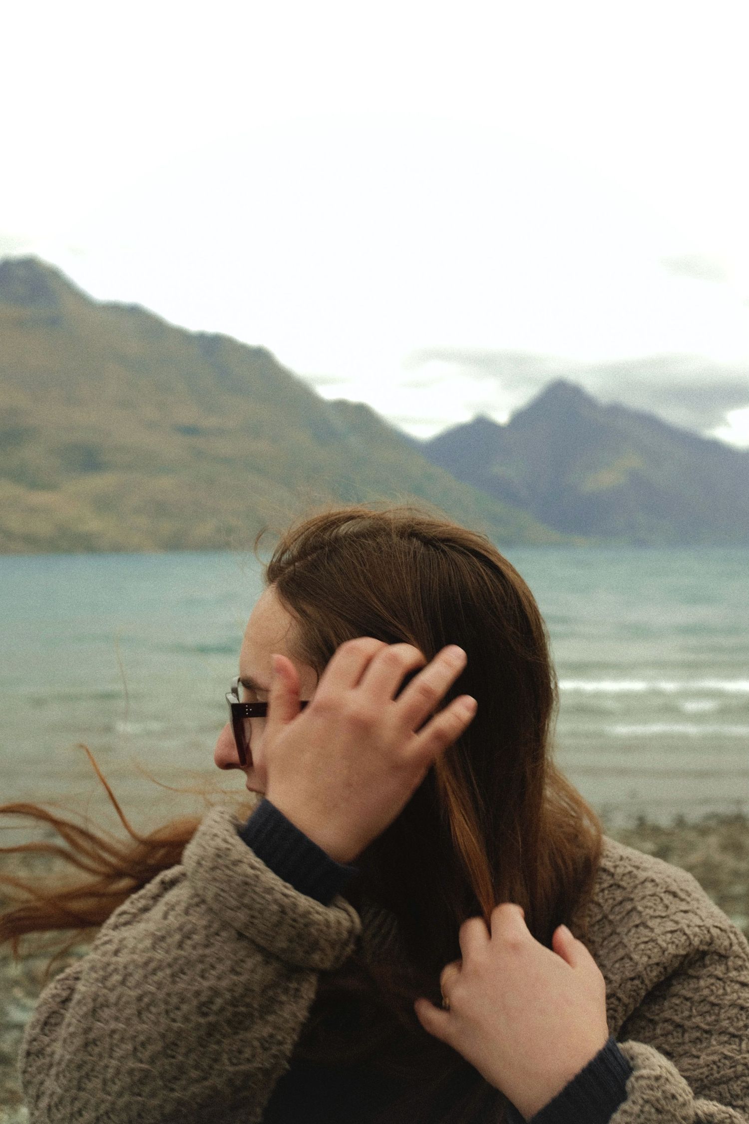 Person wearing a cozy sweater looks out at dramatic coastal mountains and ocean landscape on a cloudy day.