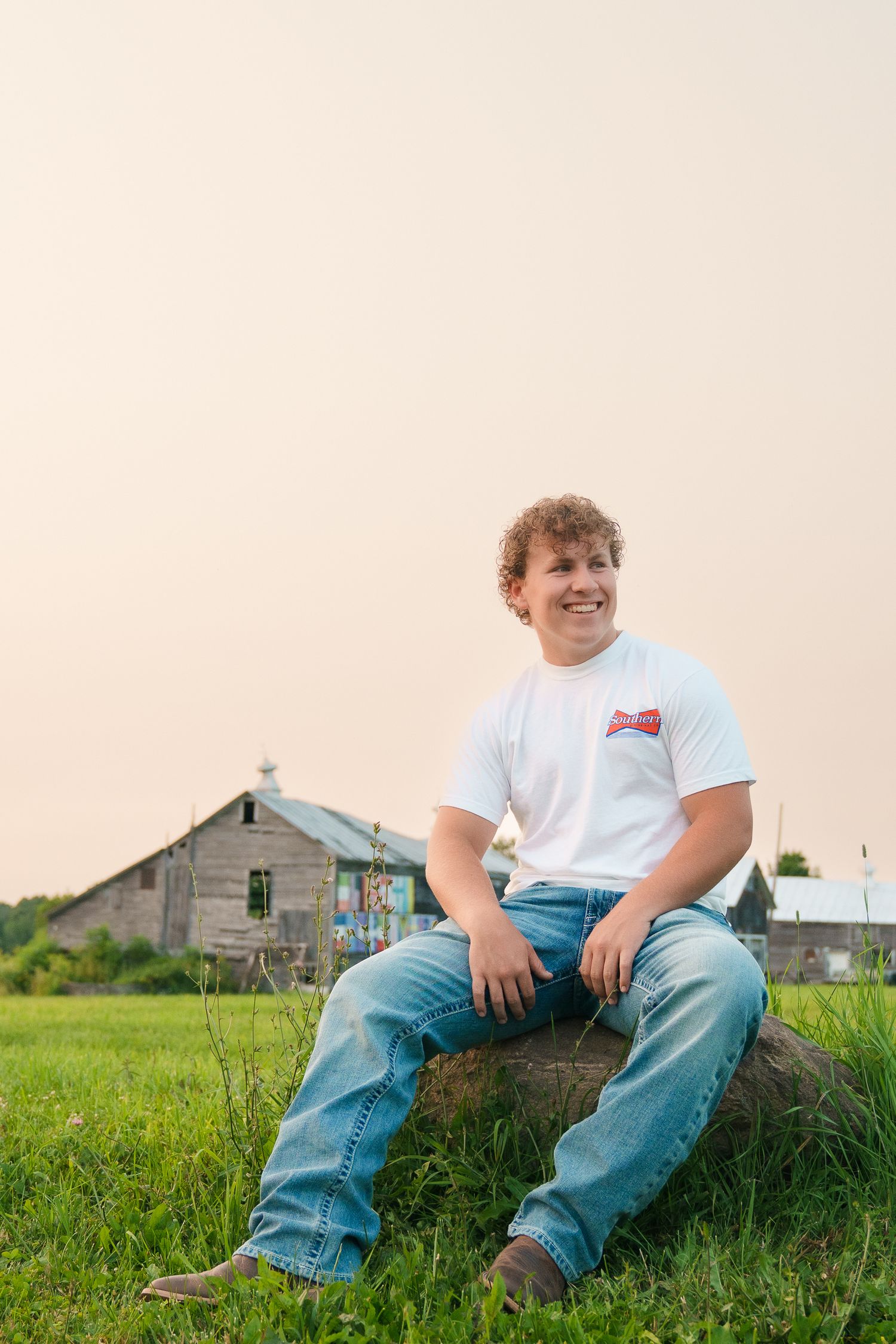 A person sits casually on grass wearing jeans and a white t-shirt with a rustic building in the background at sunset.