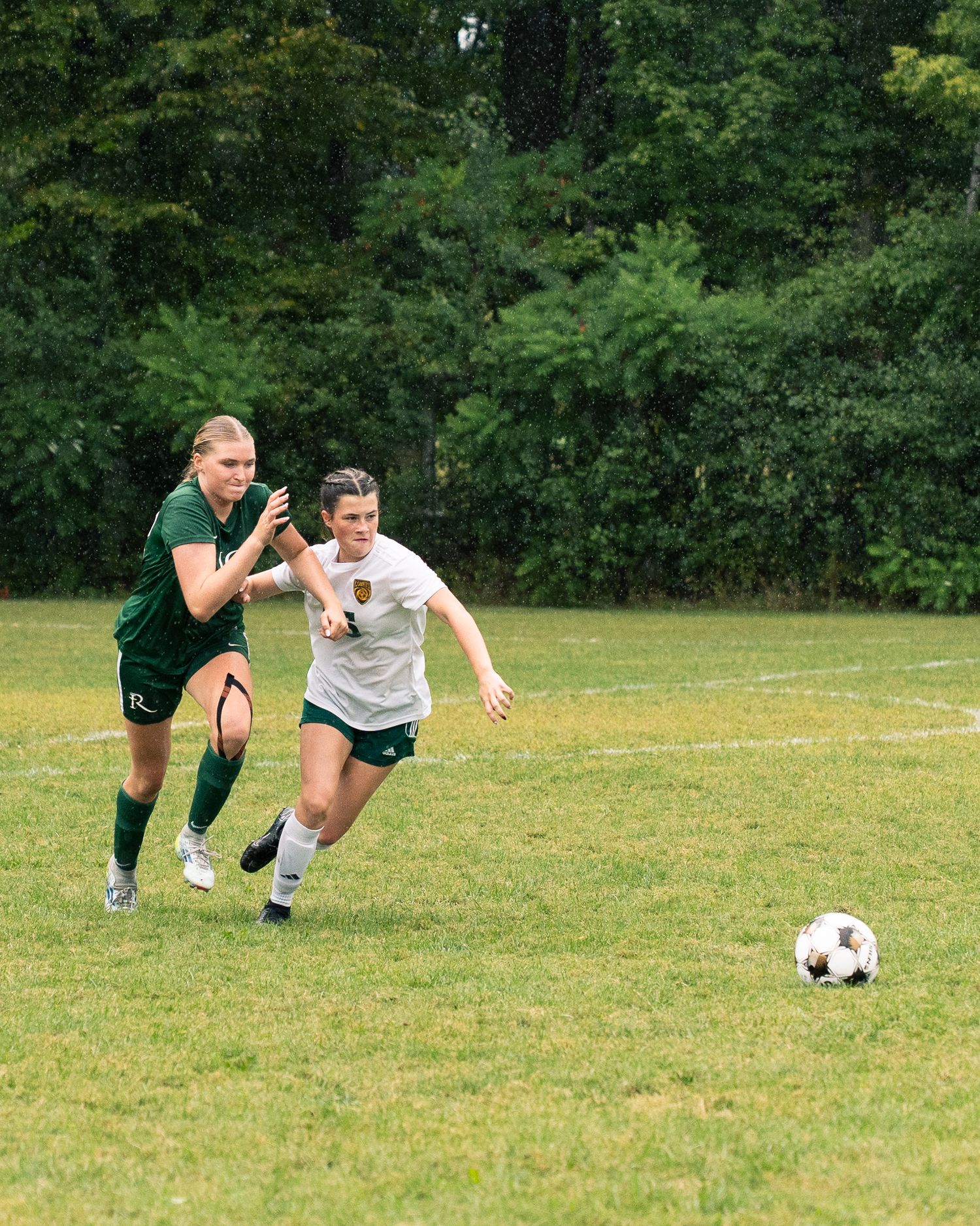 Two soccer players chase after a ball on a grassy field with trees in the background during a match.