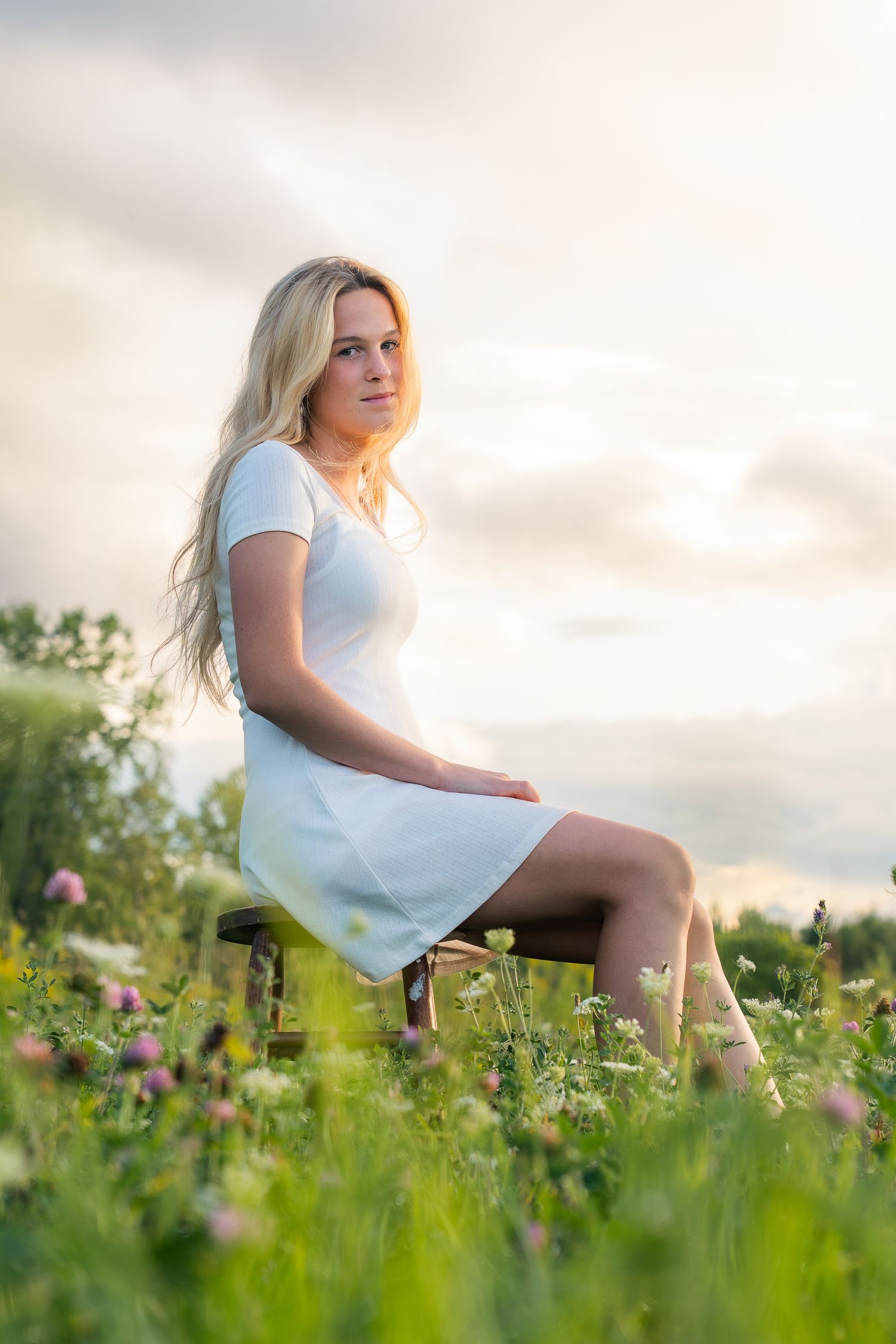 A woman in a white dress sits gracefully in a summer meadow filled with blooming wildflowers at sunset.