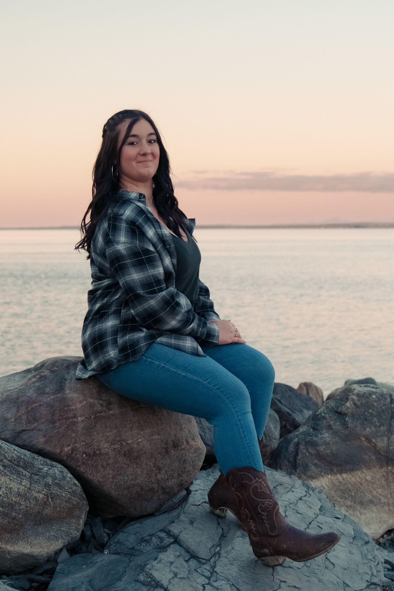 Person in plaid shirt and jeans sits on coastal rocks at sunset overlooking calm ocean waters.
