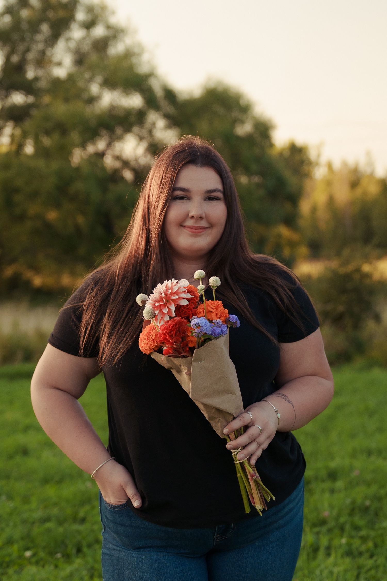 Person in black shirt and jeans holds colorful bouquet with orange and purple flowers in outdoor setting at sunset.
