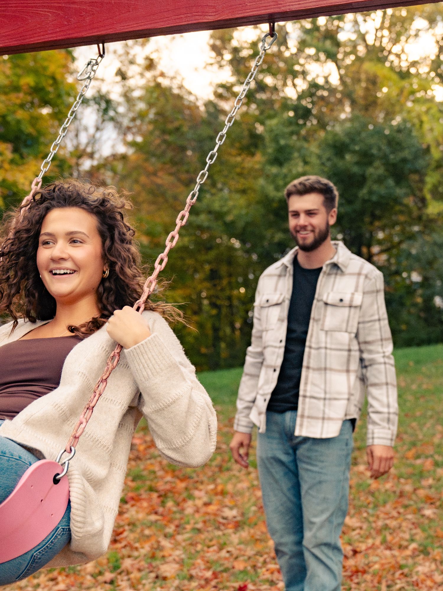 A couple enjoys a playful autumn photoshoot in a park with fallen leaves and trees in the background.