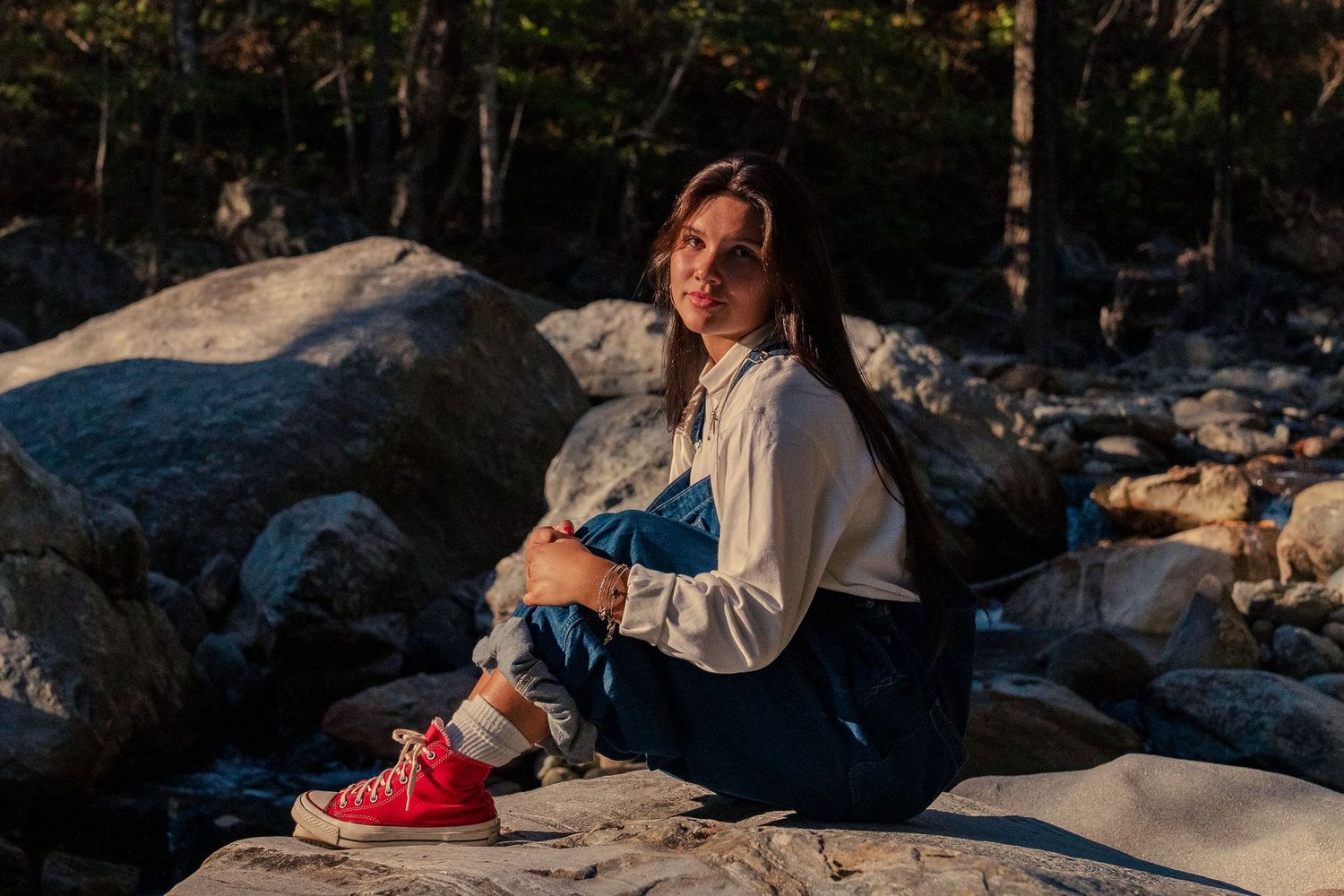 A person in a white sweater and red sneakers sits on large rocks during golden hour at sunset.