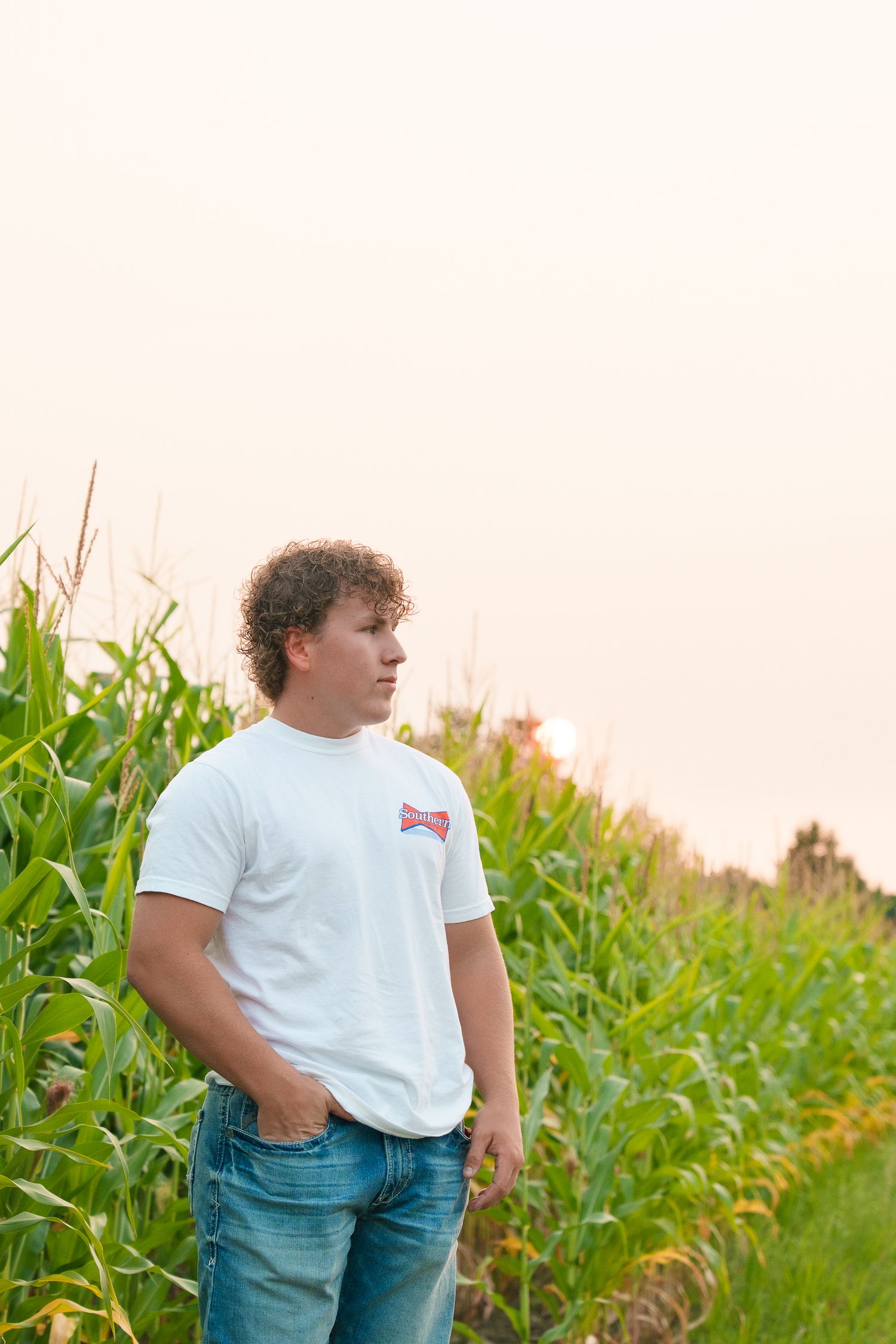 A person stands by a tall cornfield wearing a white t-shirt and jeans at dusk.