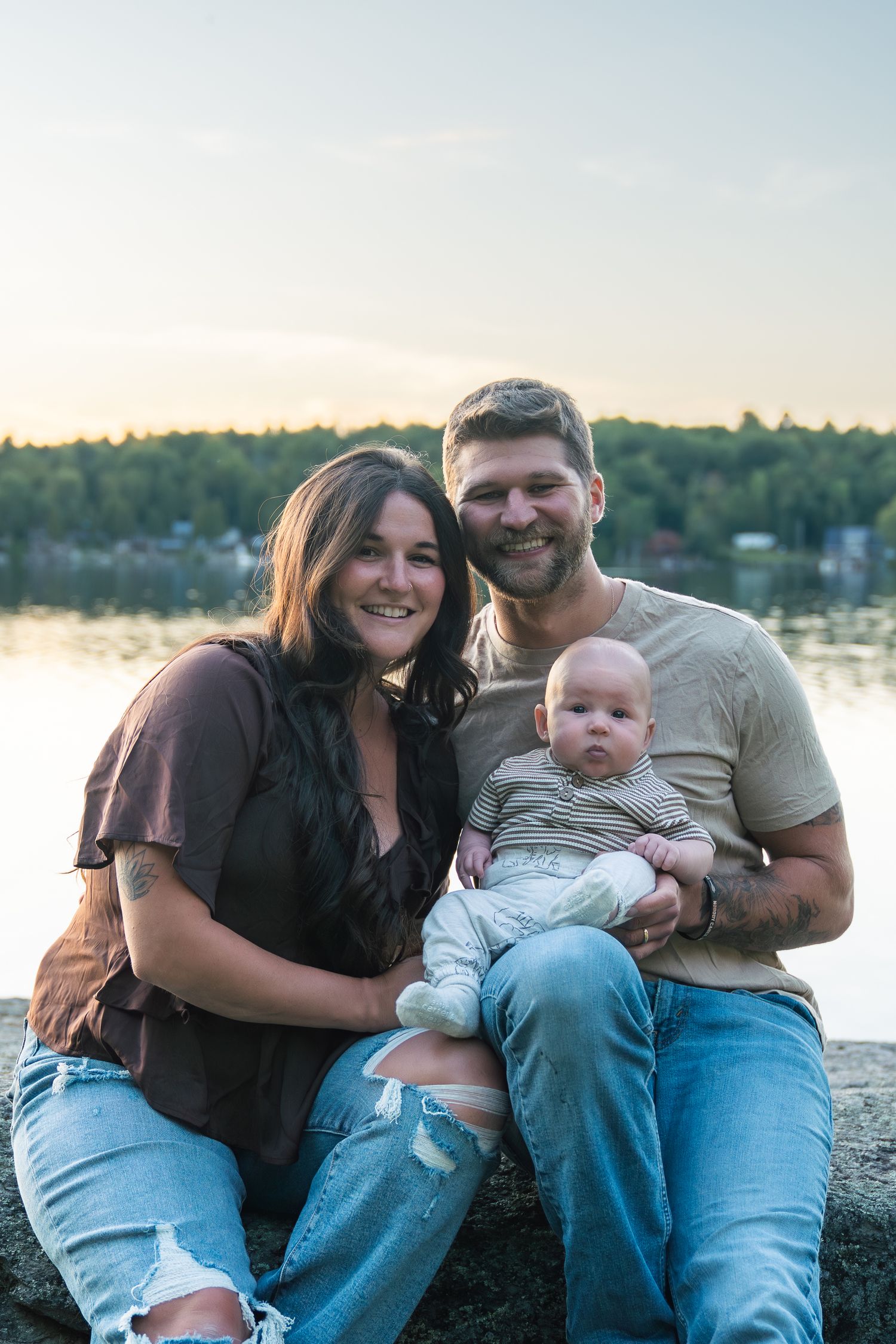 A family relaxes together on a wooded shoreline at sunset with water and trees visible in the background.