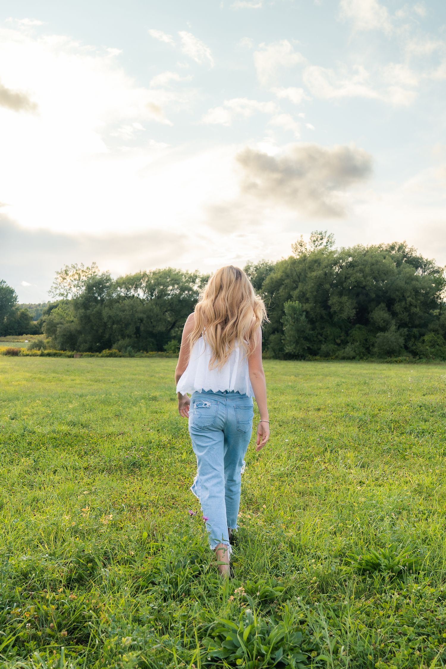 Person in light top and blue jeans walks through sunny green meadow with treeline and soft clouds in background.