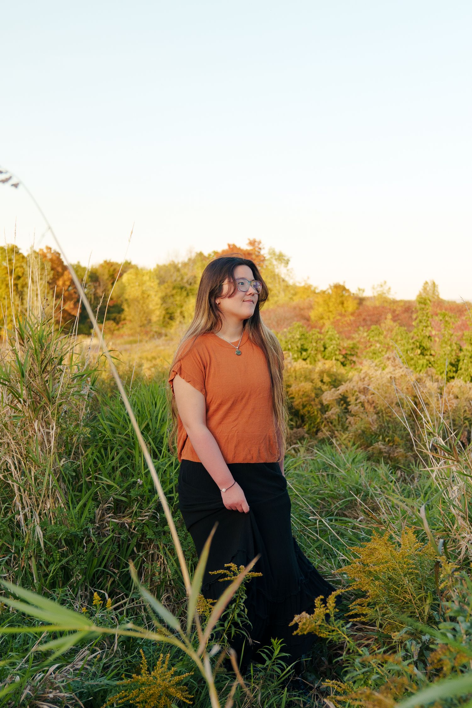 A person in an orange top and black skirt stands among tall grasses and wildflowers in a golden autumn field at sunset.