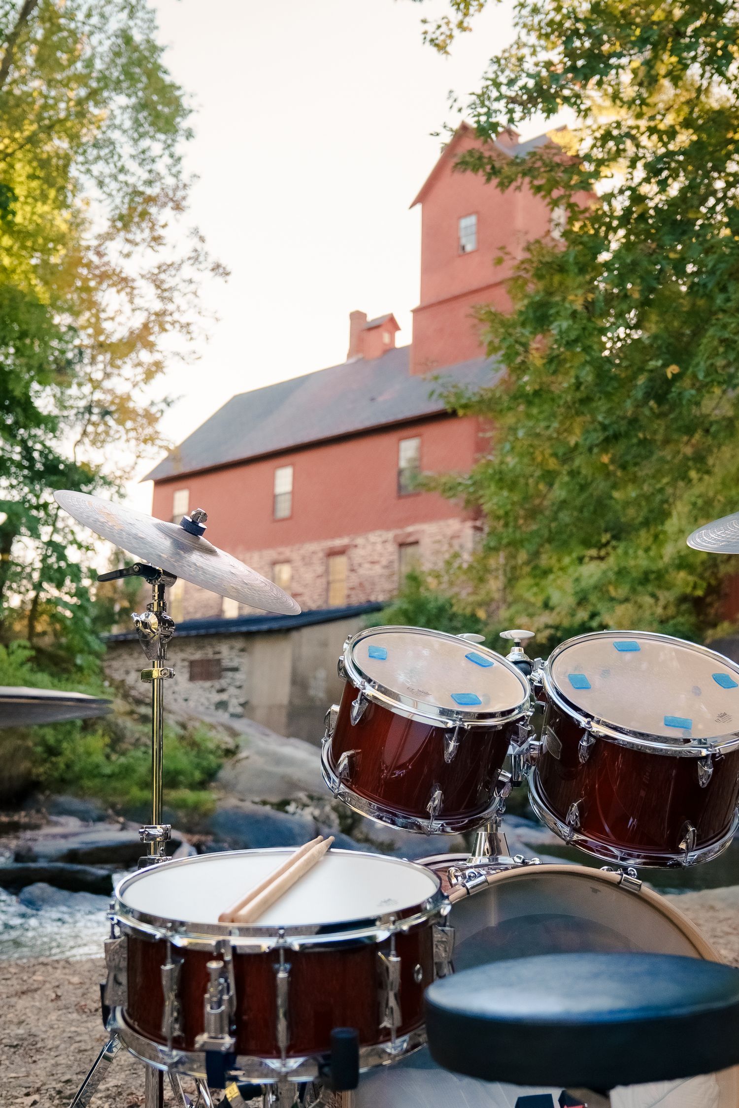 A series of drum kit images showing brown drums and cymbals arranged in a row with trees and buildings in the background.