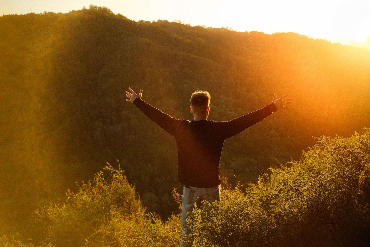 Man standing on glowing cliffside at sunset or golden hour with his back turned to the camera and outstretched arms reaching toward the sun