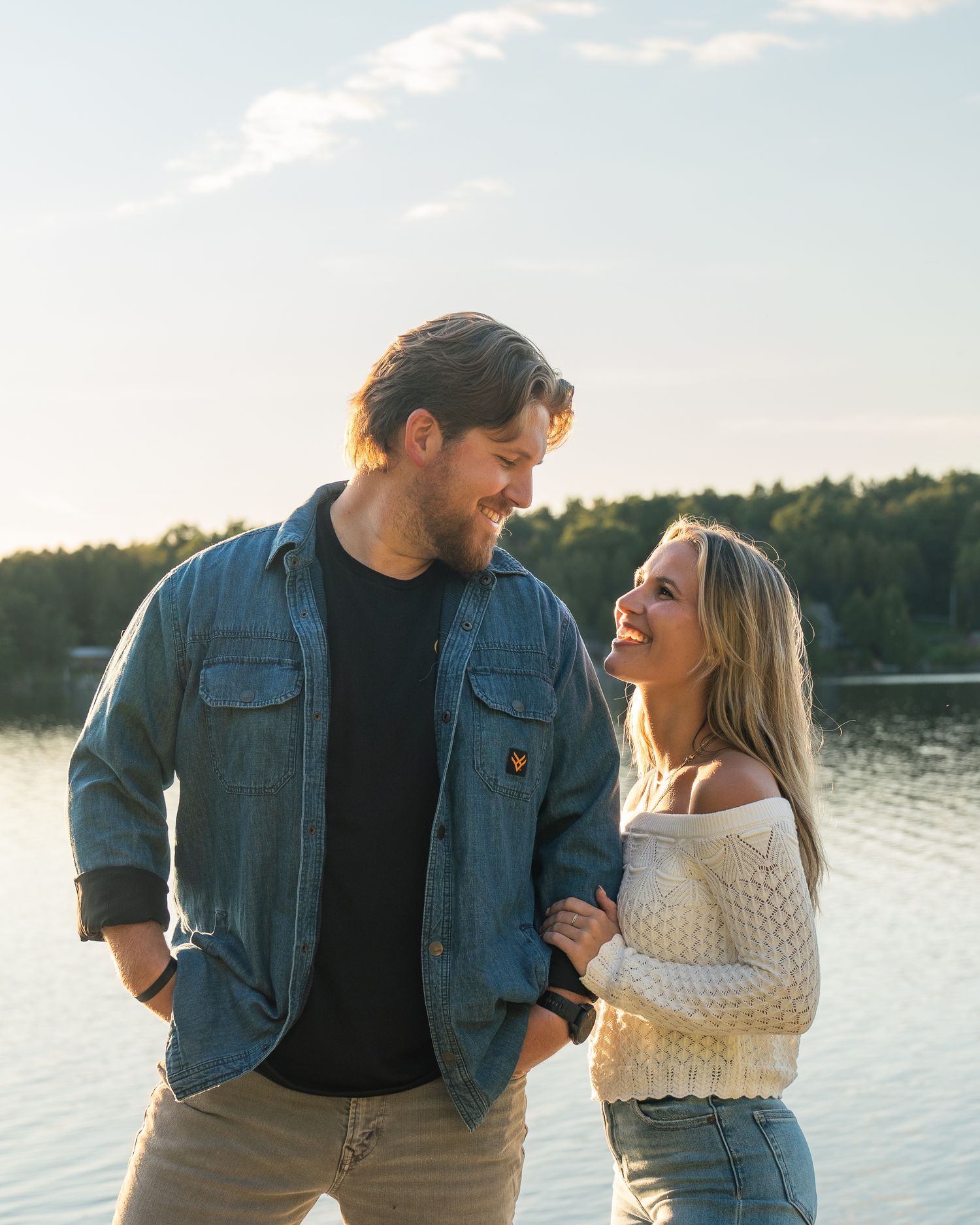 A couple stands together by a serene lake at sunset, sharing intimate moments in casual denim and white attire.