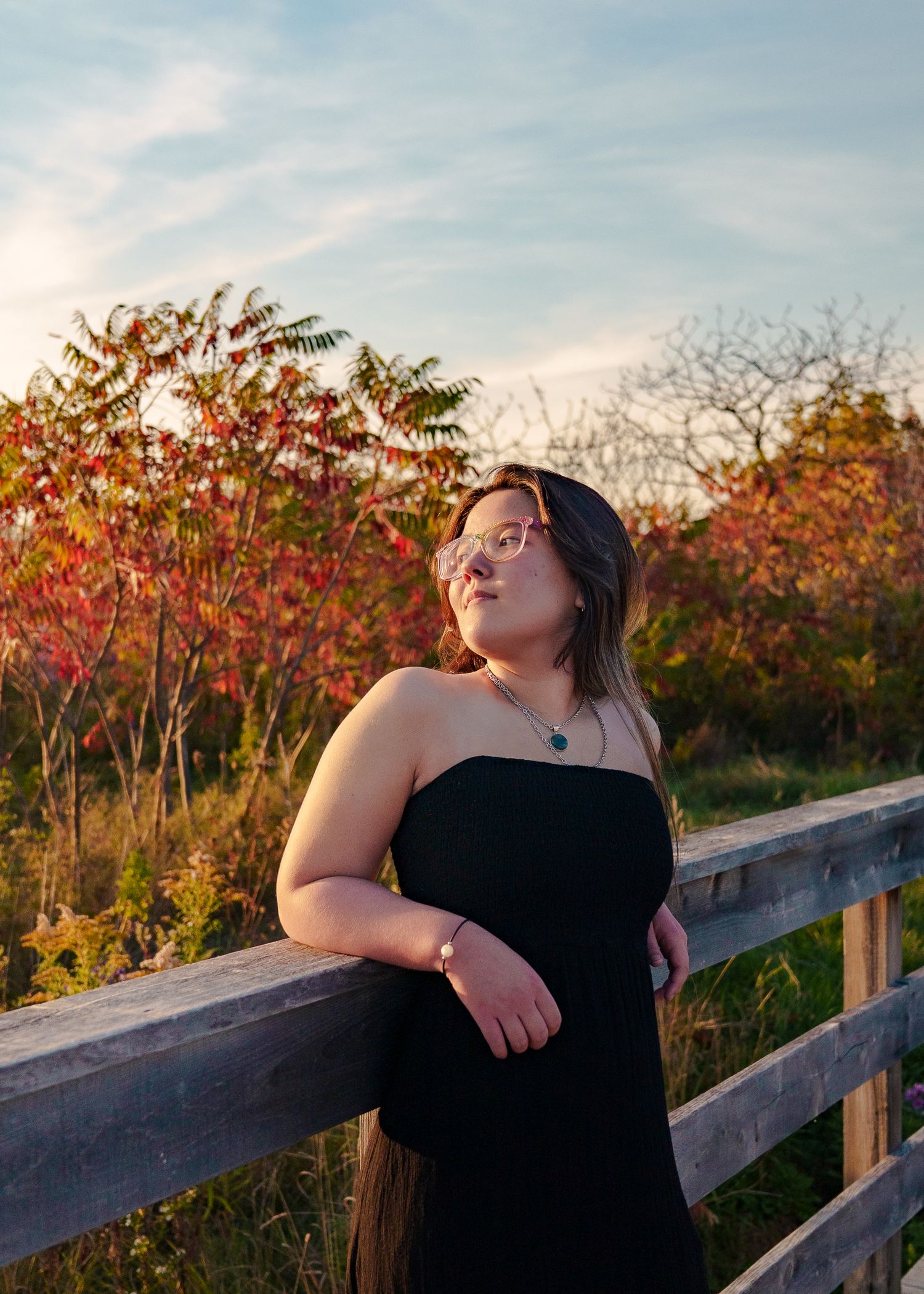 Person in black dress poses against wooden railing during sunset with vibrant red autumn foliage in background.