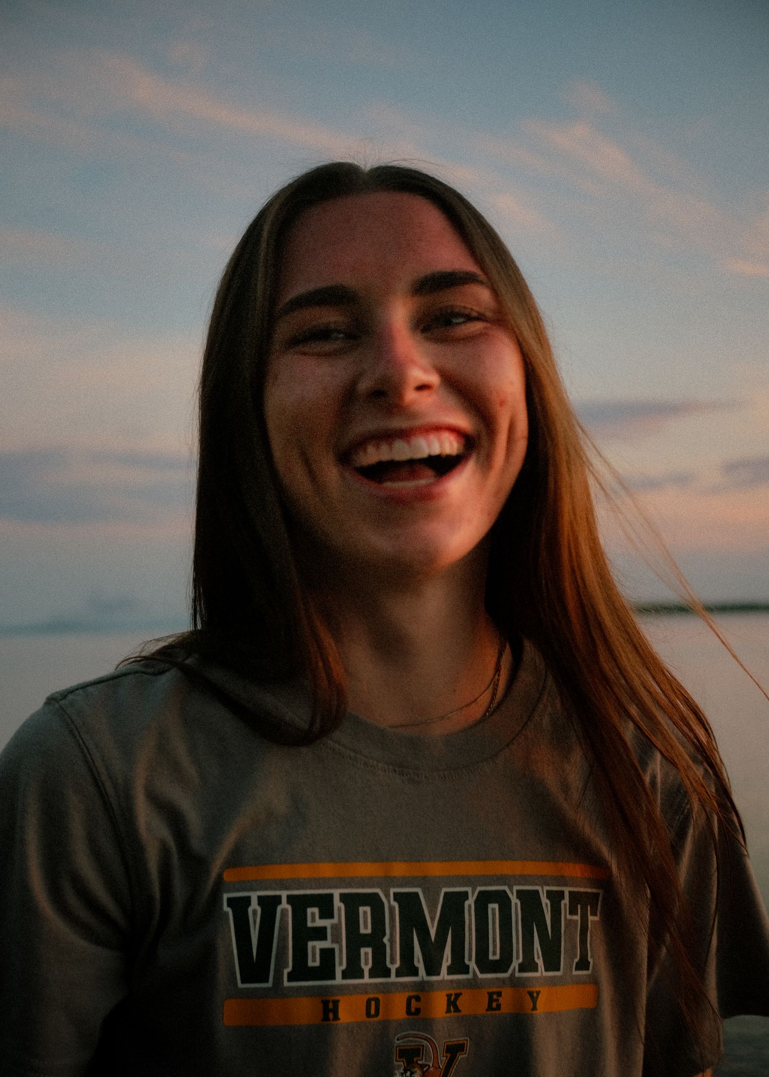 A person wearing a Vermont sweatshirt laughs during sunset by a lake.