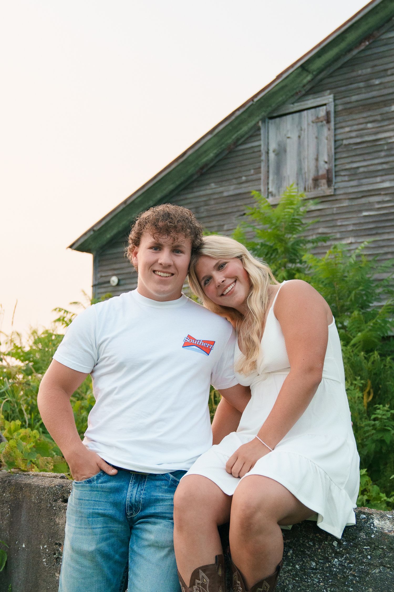 Couple posing casually against rustic wooden barn wall.
