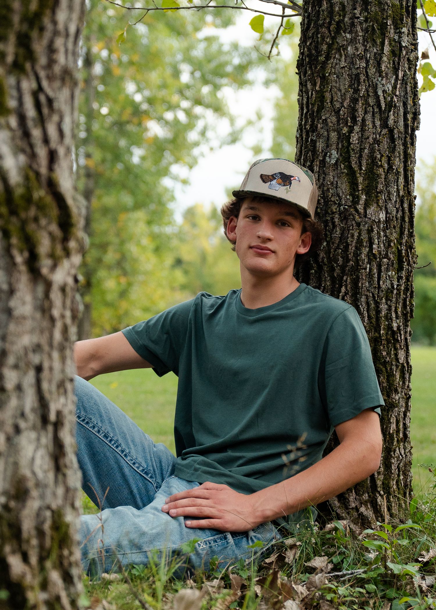 A person in a green t-shirt and baseball cap relaxes against a tree trunk in a natural outdoor setting.