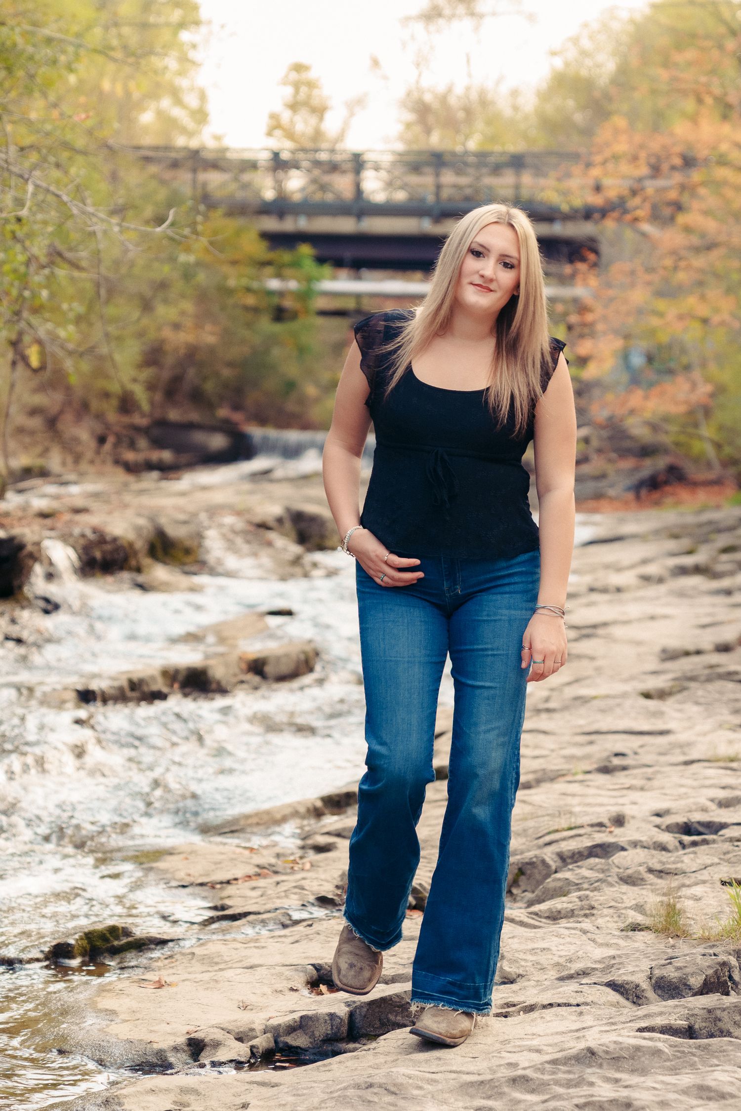 Person in blue jeans poses on rocky riverbank with autumn trees and bridge in background.