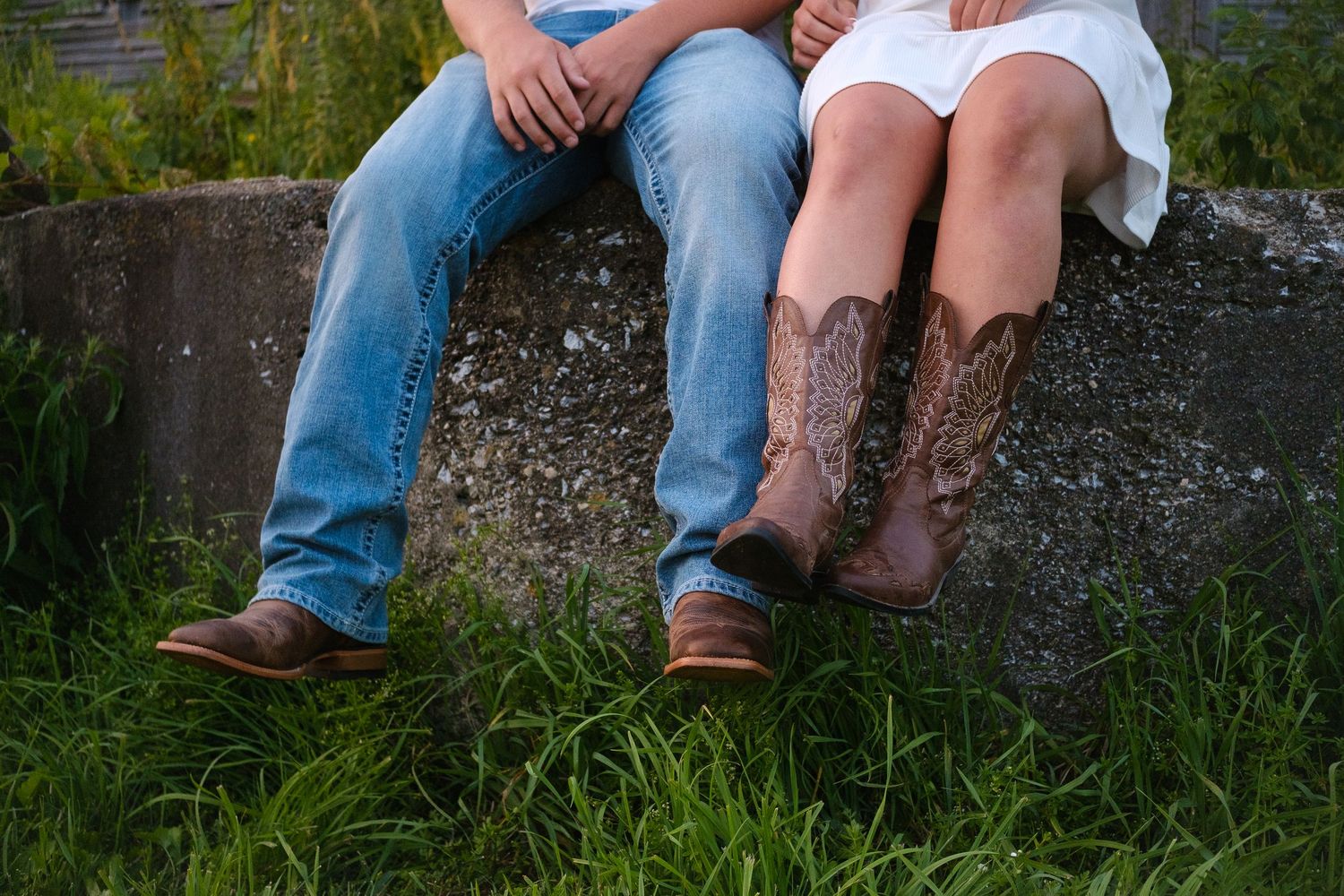 Feet of a couple sitting together on a concrete wall surrounded by grass and nature.